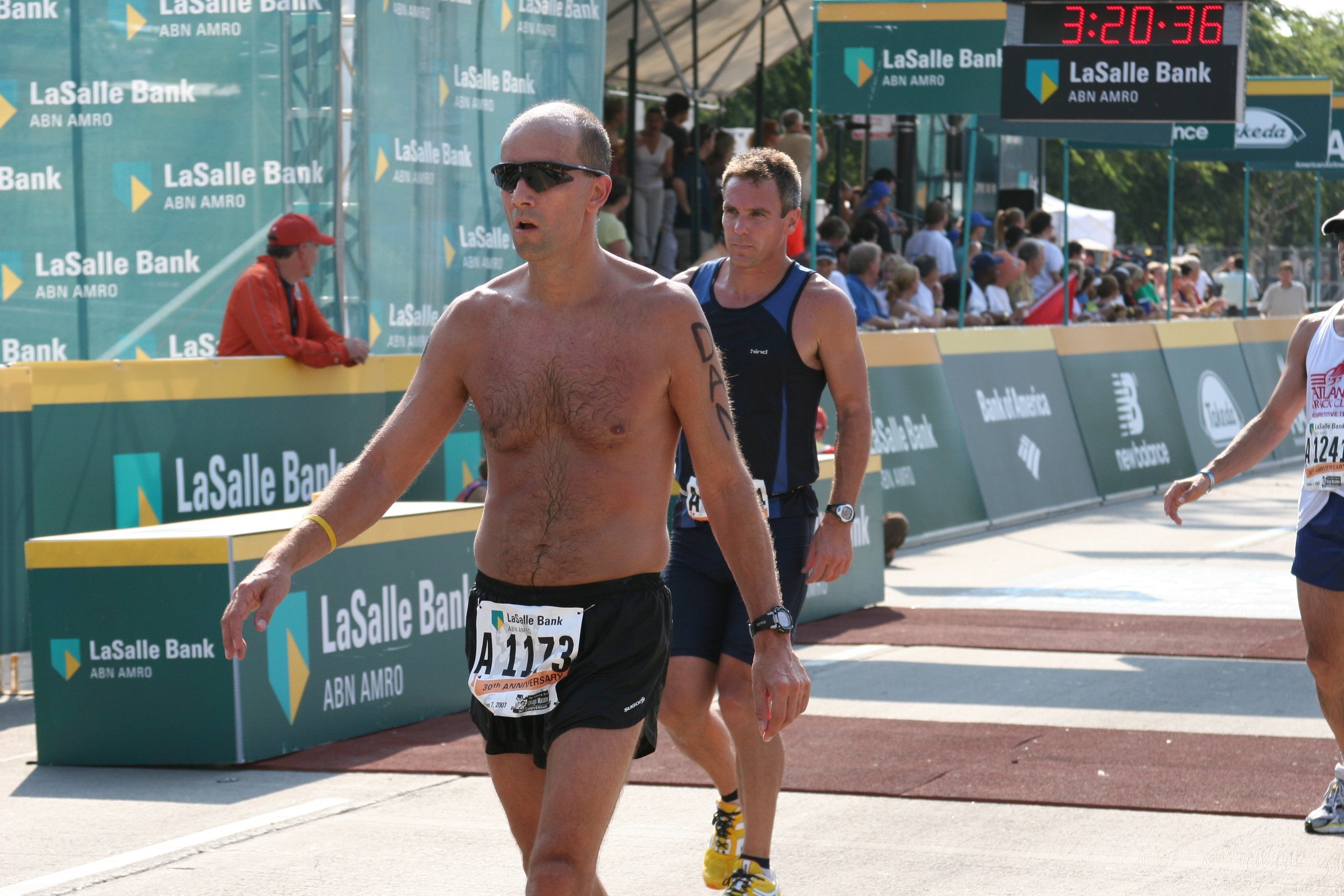 Male marathon runner with sunglasses, shirtless, and a bib number A1173 walking near the finish line during a race, with spectators and a digital clock showing 3:20:36 in the background.