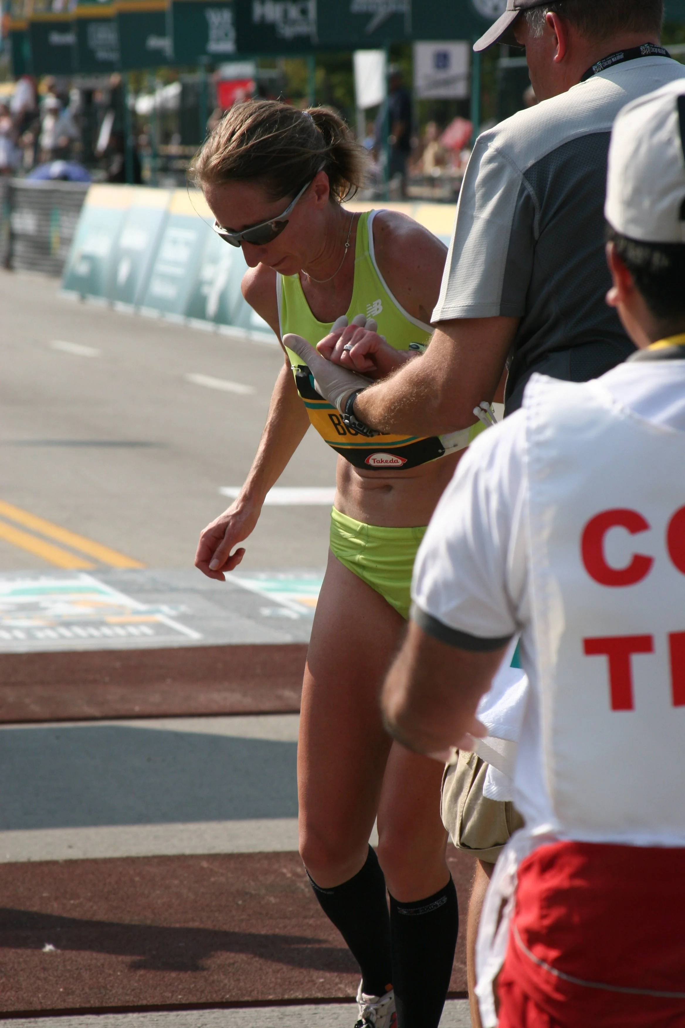 A female marathon runner leaning forward during a race, checking her watch with support from personnel on the track, with spectators and event banners in the background.