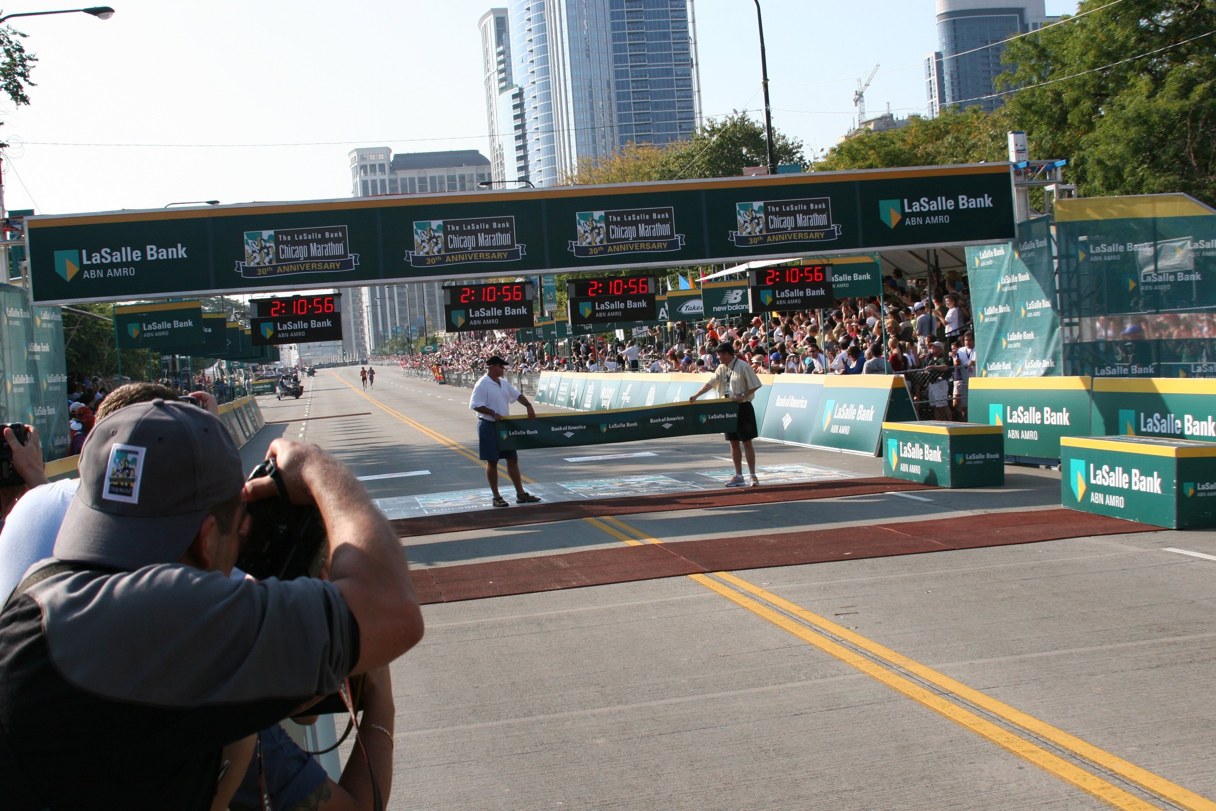 Start line of the Chicago Marathon with a large crowd of runners and spectators, digital clocks, and sponsor banners, with tall city buildings in the background.
