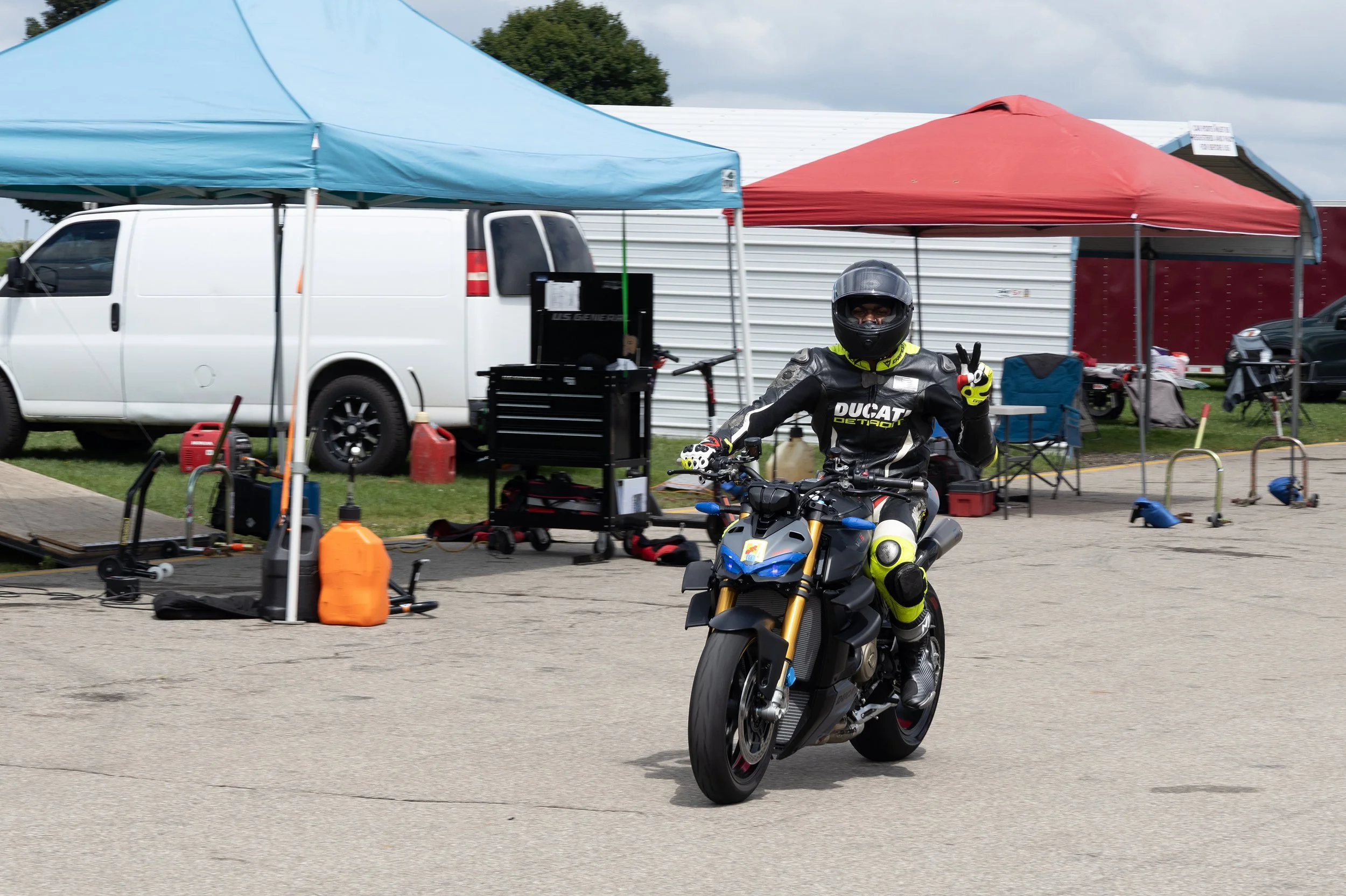 A motorcyclist in black leather gear and a helmet, sitting on a black and blue motorcycle, is making a peace sign with their right hand at an outdoor event. There are tents, equipment, and vehicles in the background.