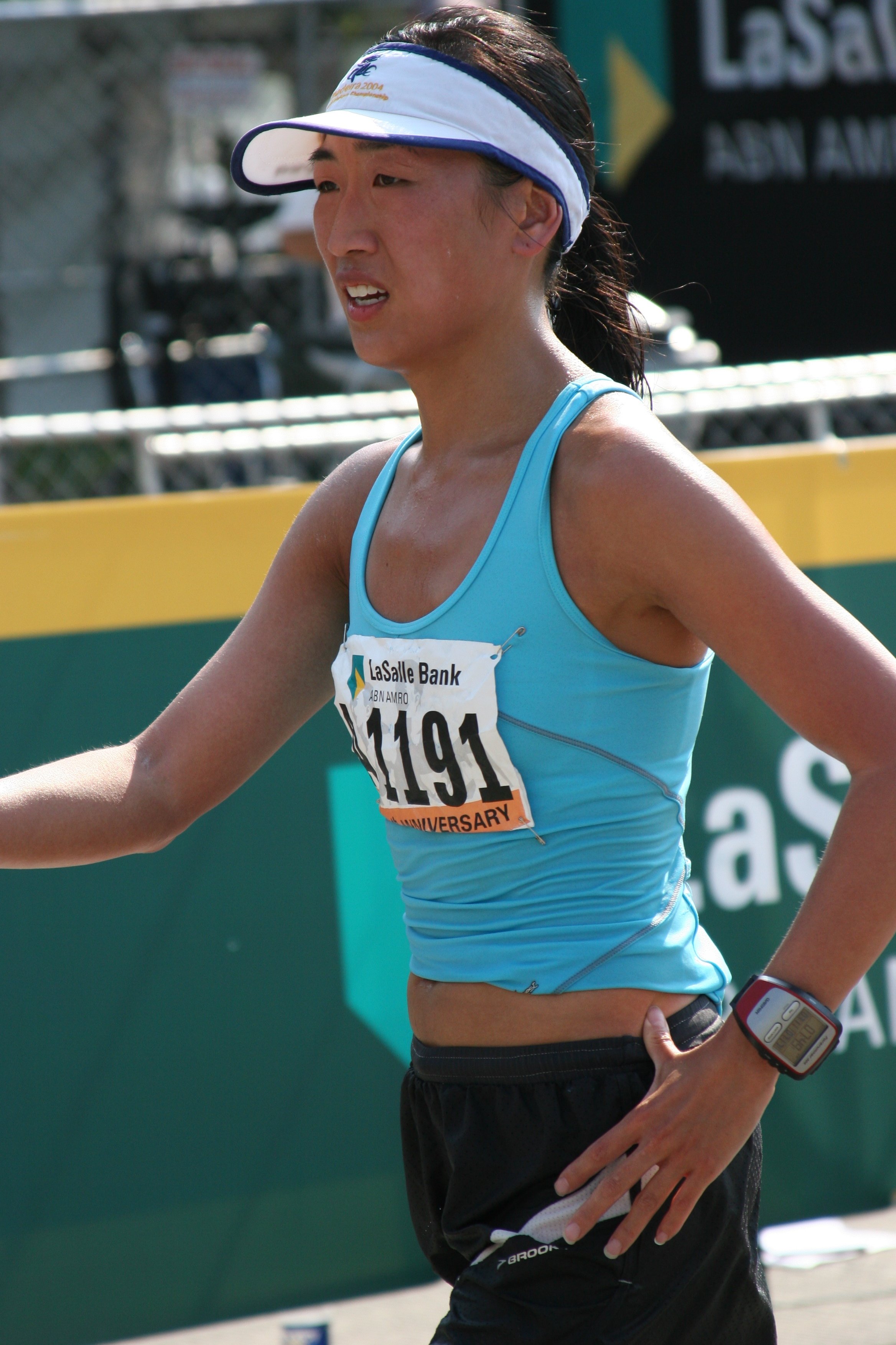 A female marathon runner wearing a blue tank top, a white visor, a race bib with the number 1191, and a sports watch on her left wrist, running outdoors during the daytime.