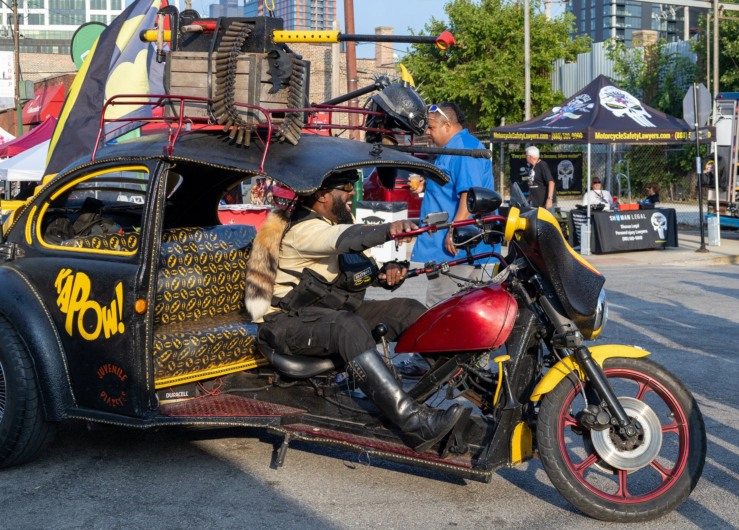 A man riding a red motorcycle with a sidecar decorated with Batman logos. The sidecar has a spiked helmet and a Batman-themed seat cover. In the background, there is a man in a blue shirt and sunglasses, standing near tents and booths at an outdoor e