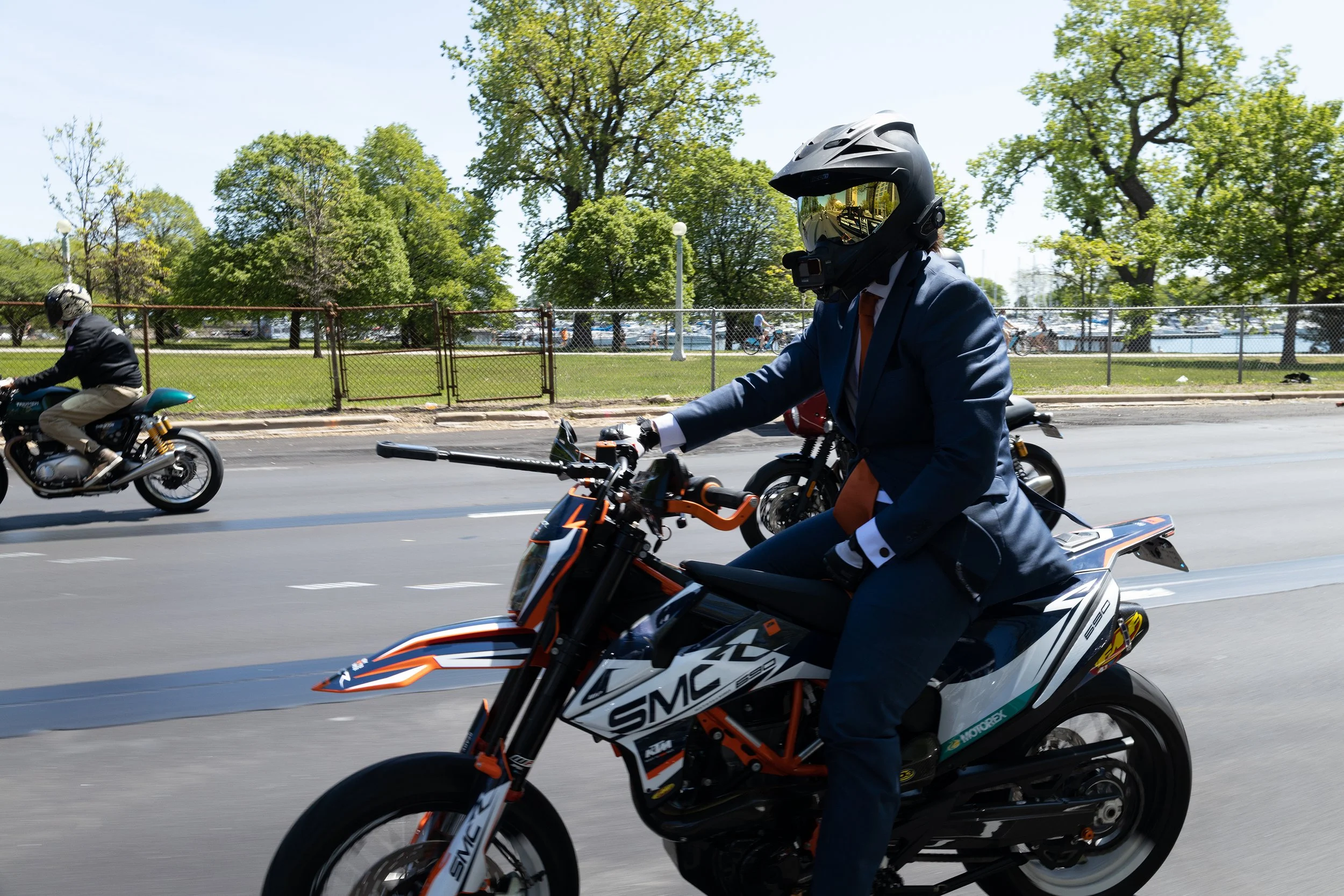 A man in a business suit riding a motorcycle on a street, wearing a motorcycle helmet. In the background, there are trees, a fence, and park with people riding bicycles.