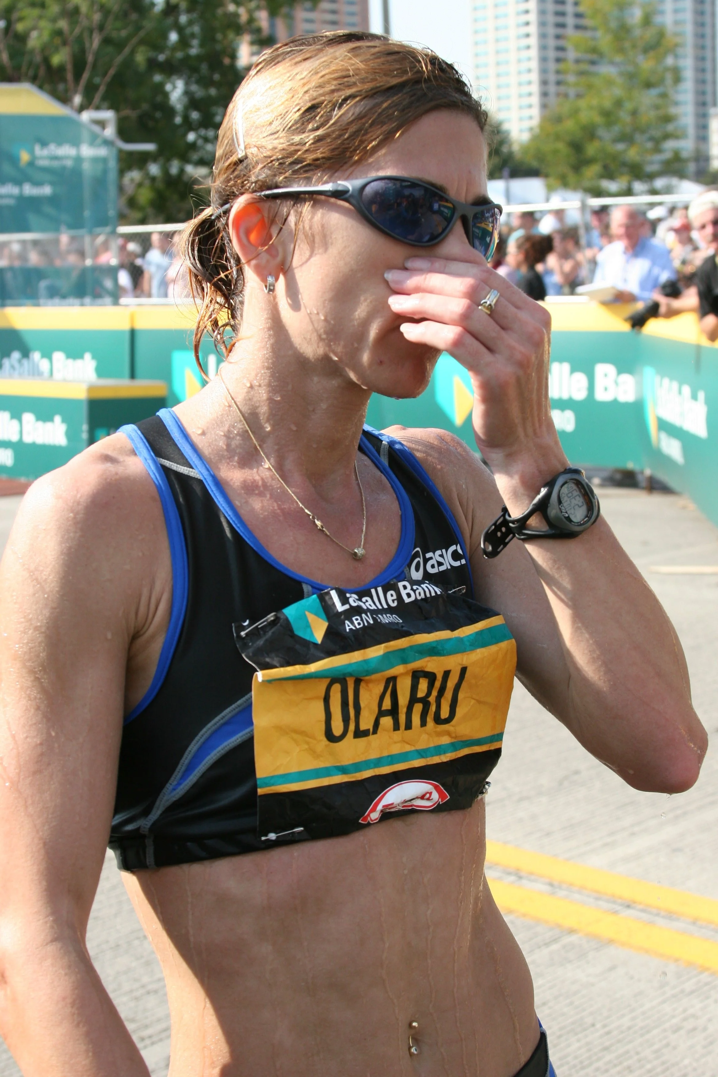 A woman in running attire, wearing sunglasses and a race bib with the name 'Olaru', appears to be emotional or exhausted after a race, with her hand covering her face. There are spectators and event signs in the background.