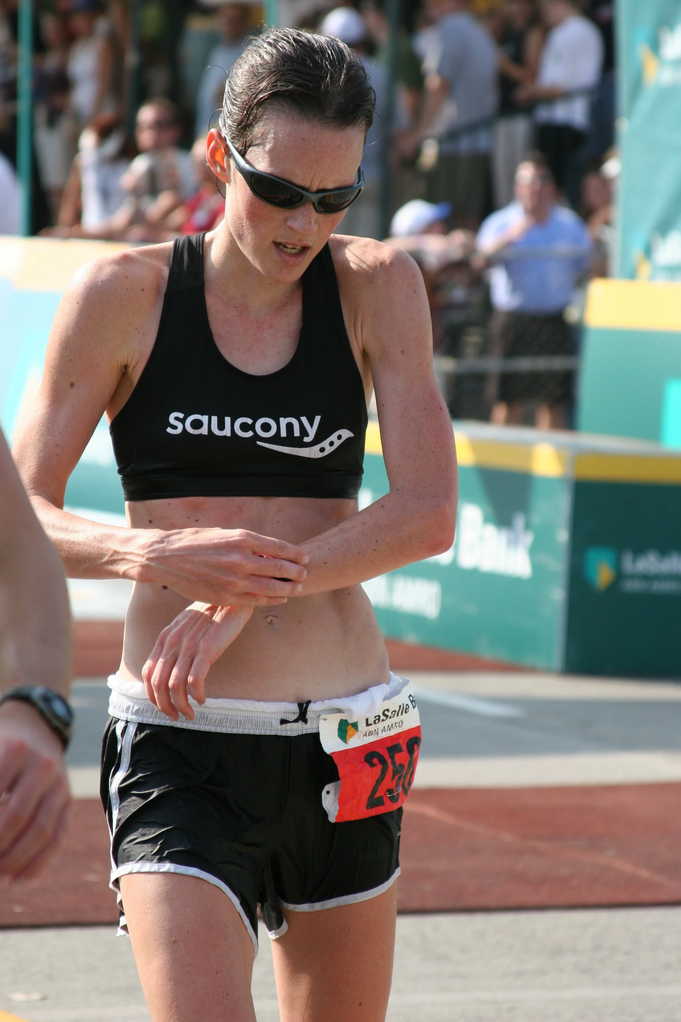 A female marathon runner wearing sunglasses and a black Saucony sports bra, with a race bib numbered 250, is running outdoors as spectators watch in the background.