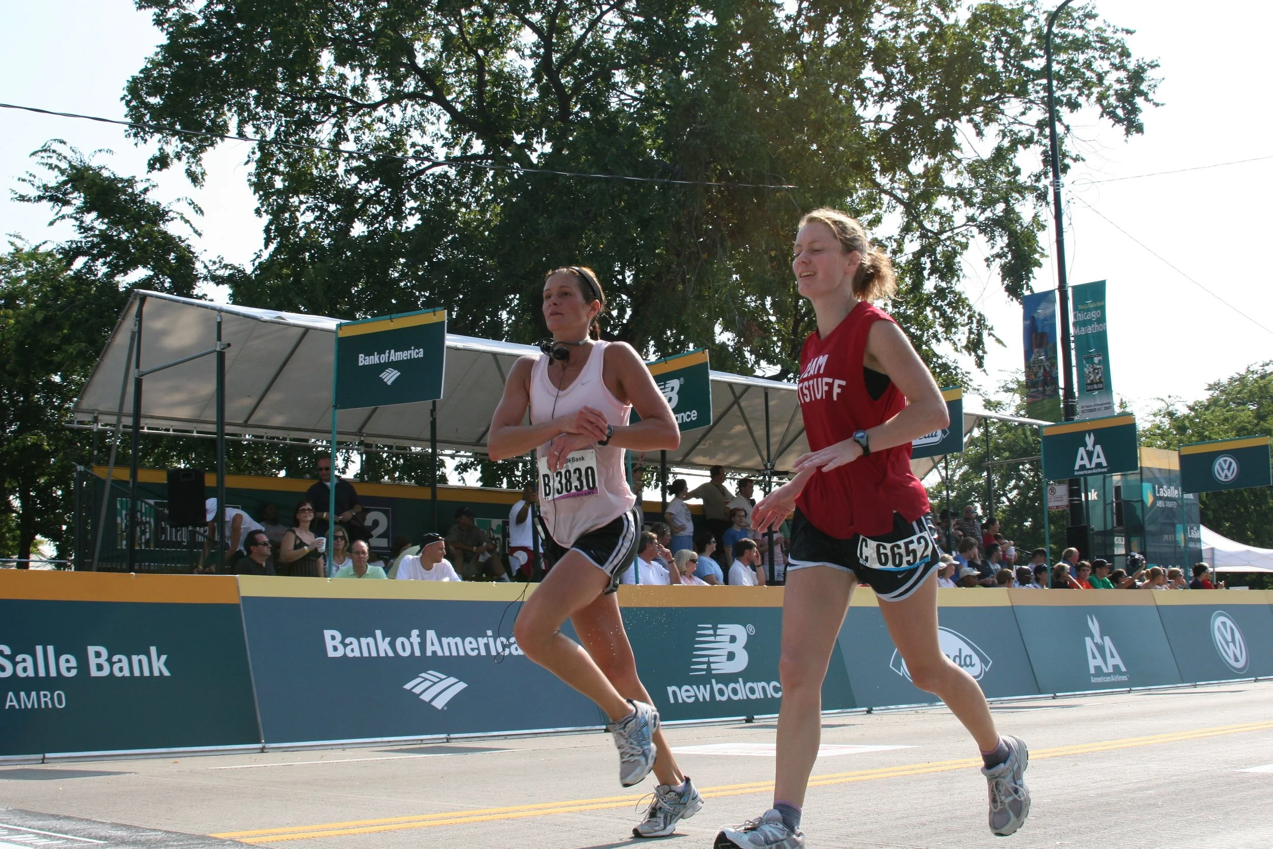 Two female marathon runners in athletic gear running on a city street near a crowd of spectators and a finish line with banners and sponsorships.