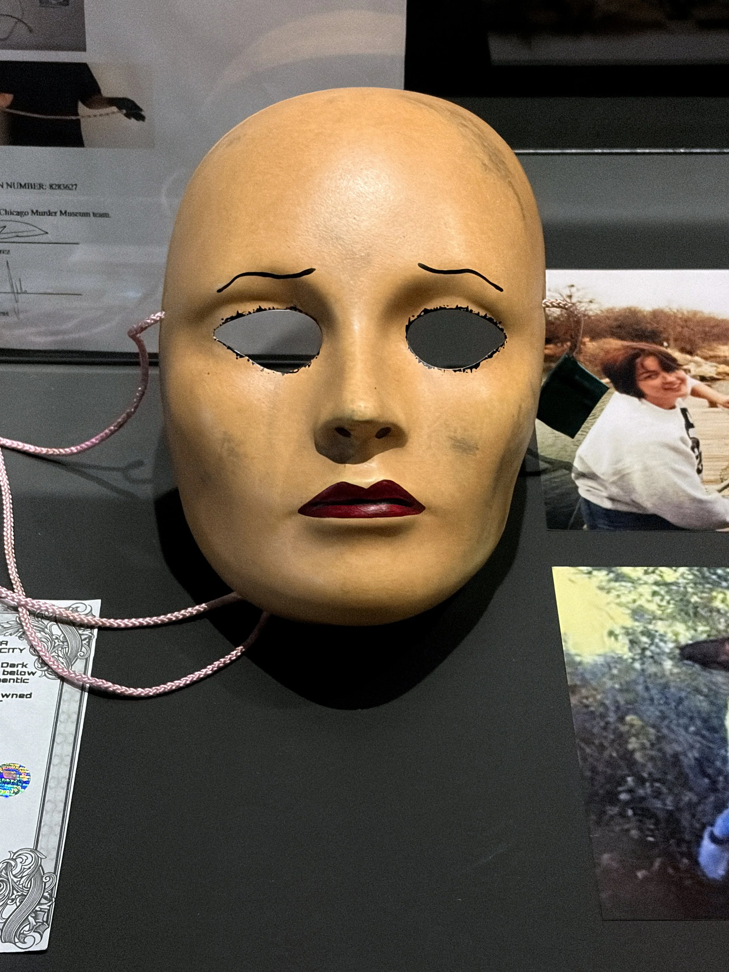A vintage lady's mask with a painted face, black eye holes, red painted lips, and drawn eyebrows, laying on a flat surface with photographs around it.