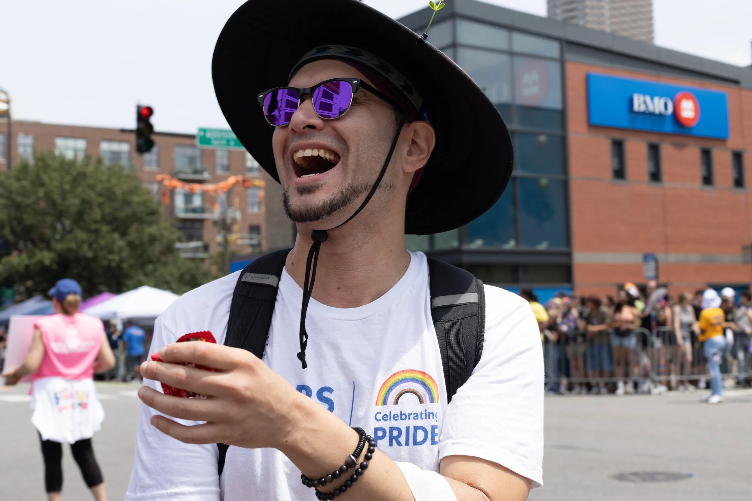 A person wearing sunglasses, a wide-brimmed hat, and a white t-shirt with a rainbow and Pride logo, smiling and holding a small red item at an outdoor LGBTQ+ pride event in an urban setting.