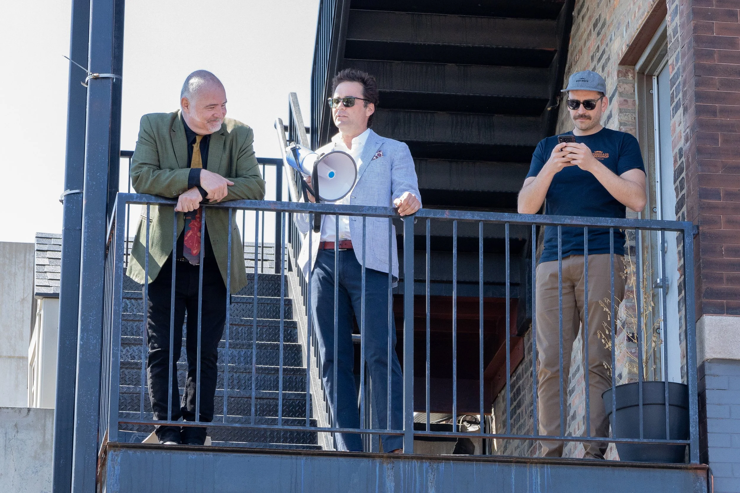 Three men standing on a small balcony outside a brick building, one looking at a phone, one with a megaphone, and one leaning on the railing.