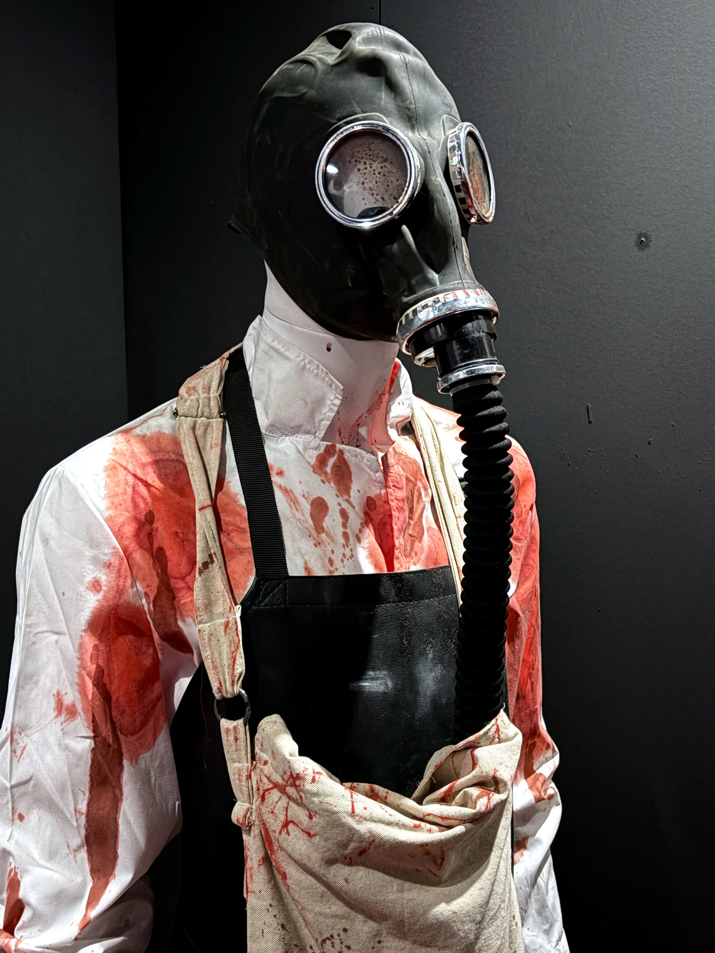 Life-sized mannequin wearing a black gas mask with a hose, blood-stained white medical gown, and harness, set against a dark background.