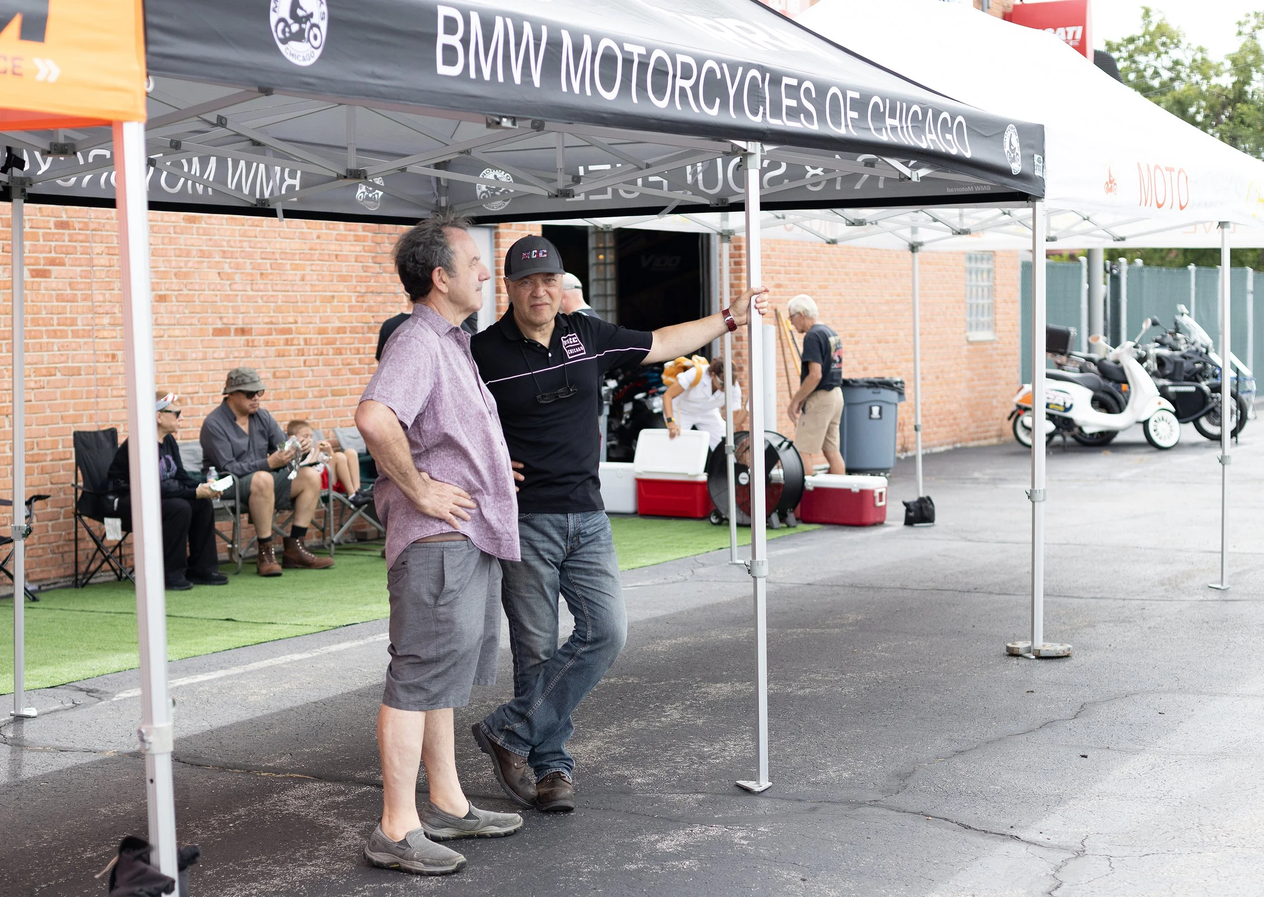 Two men are talking under a black canopy tent that reads 'BMW MOTORCYCLES OF CHICAGO'. There are several people sitting on chairs in the background, and motorcycles are parked nearby against a brick wall.