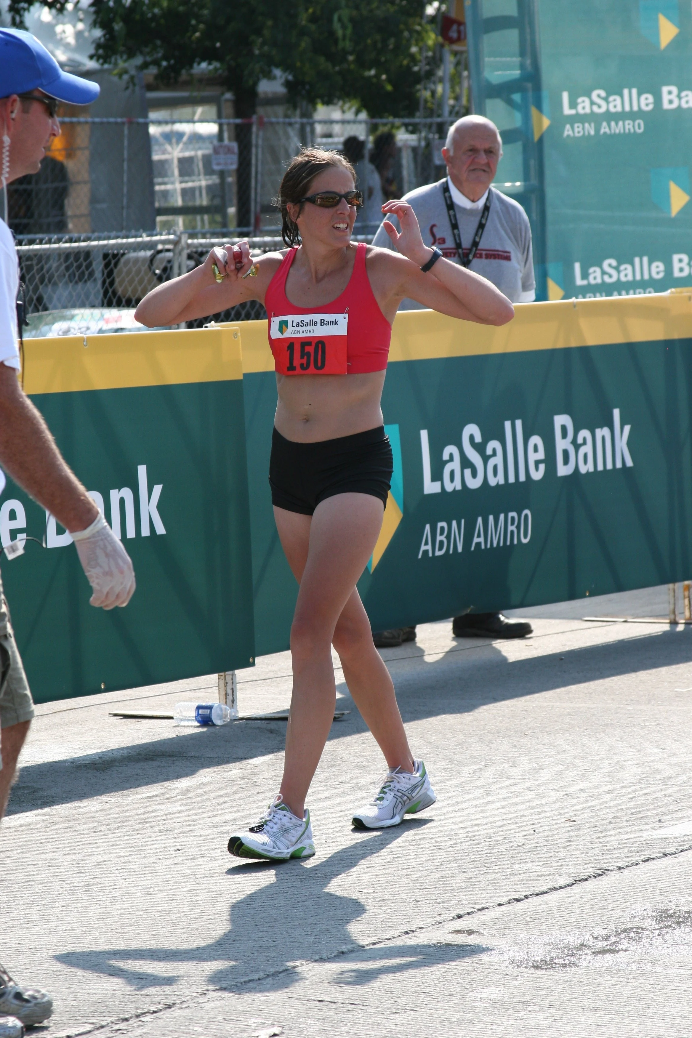A female marathon runner wearing a red sports bra and black shorts, with bib number 150, is at a race finish line. She is wearing sunglasses, white running shoes, and appears to be catching her breath or stretching. There are race officials or volunt