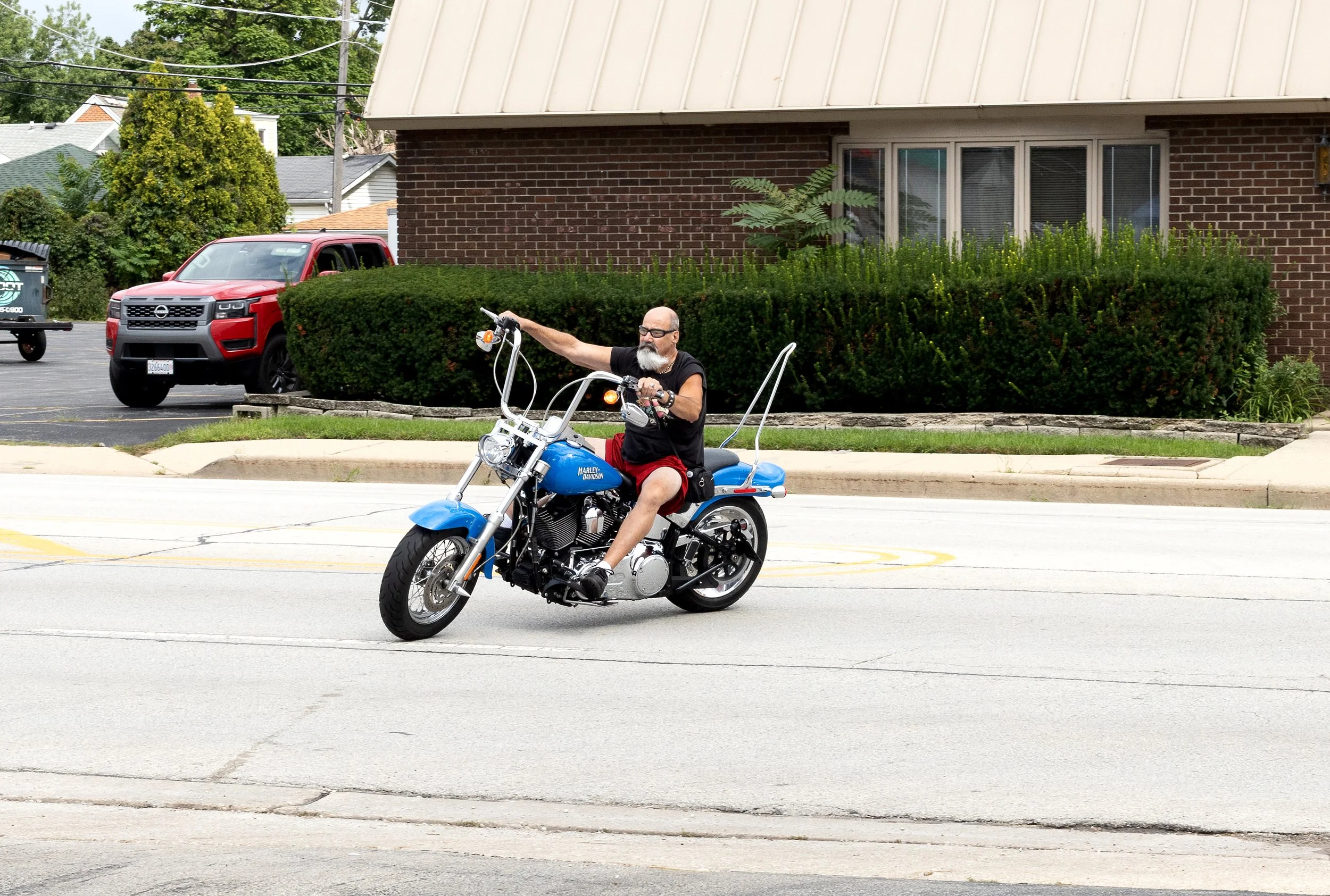 A man with a beard and glasses riding a blue Harley Davidson motorcycle on a street, with a red pickup truck and a house with bushes and windows in the background.