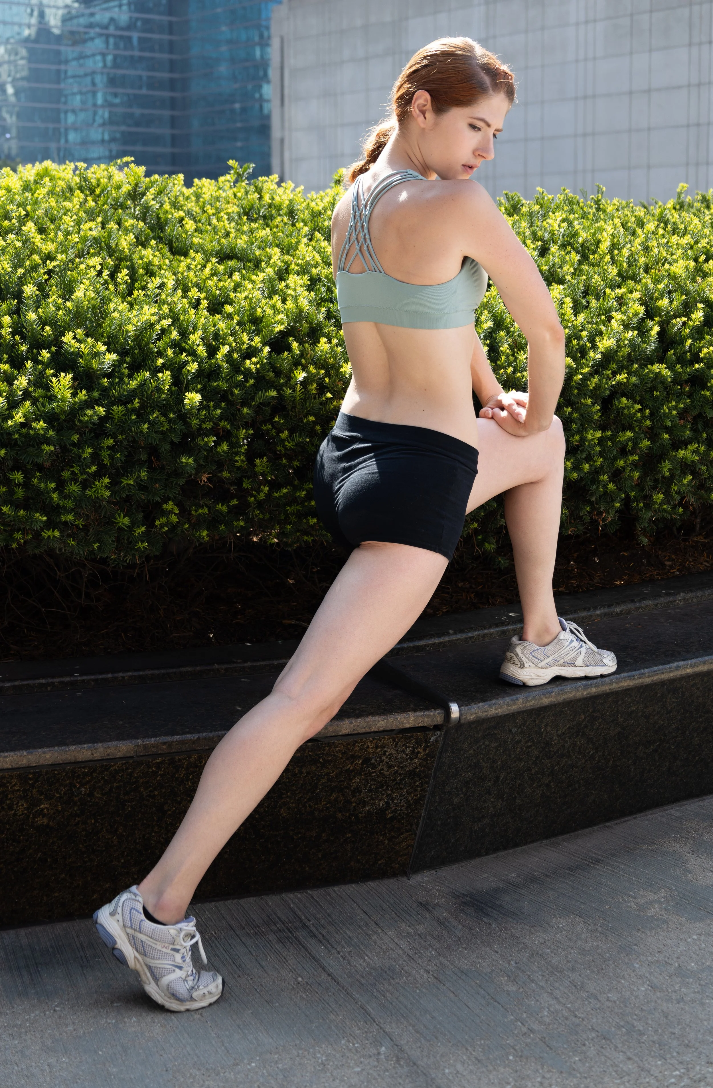 Woman in athletic clothing stretching outdoors next to a green bush and modern buildings.