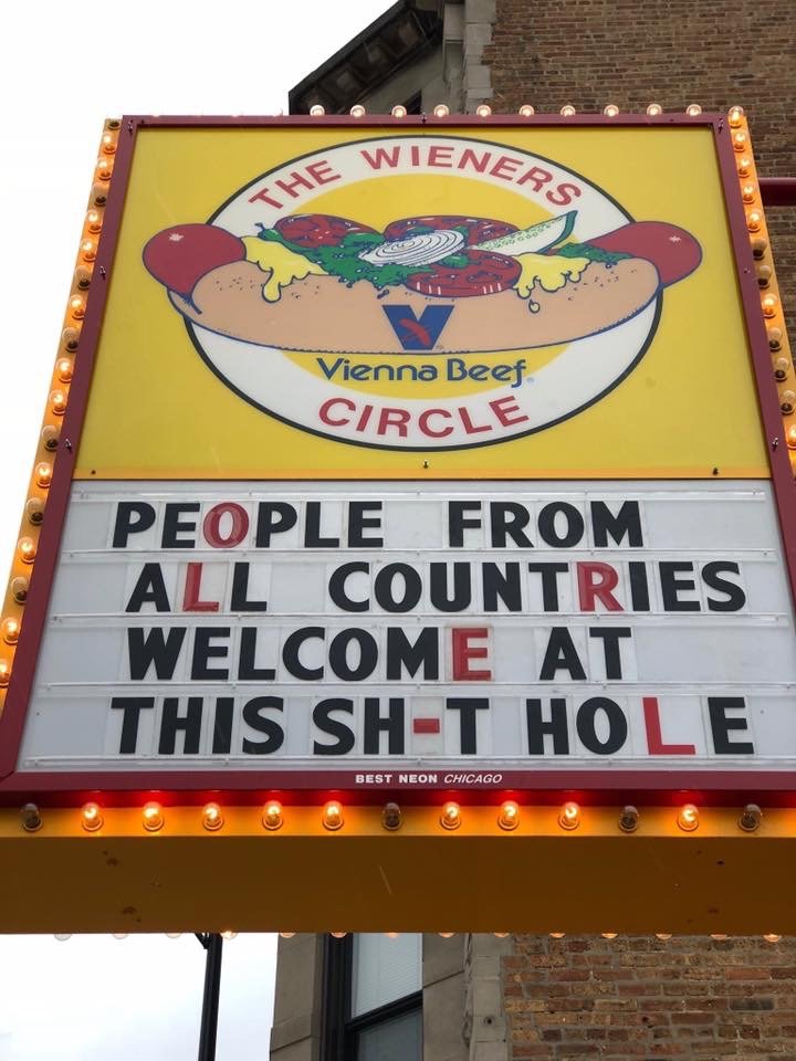 A colorful restaurant sign for 'The Wieners Circle' featuring an illustration of a hot dog with toppings. The sign also displays a humorous message welcoming people from all countries to the establishment.