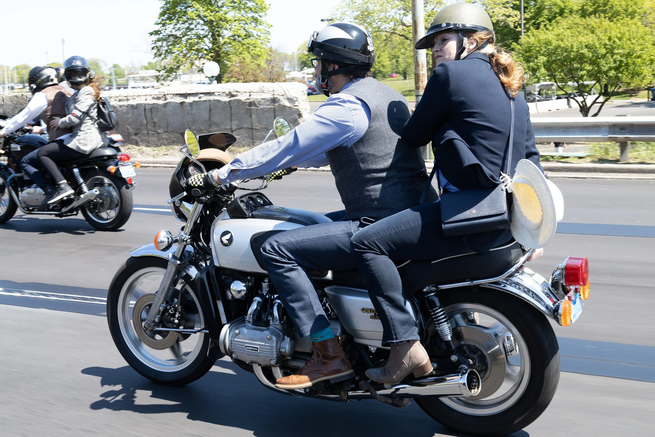 Two women riding a motorcycle, wearing helmets, with one carrying a sun hat and a purse, on a sunny day with trees and a stone wall in the background.