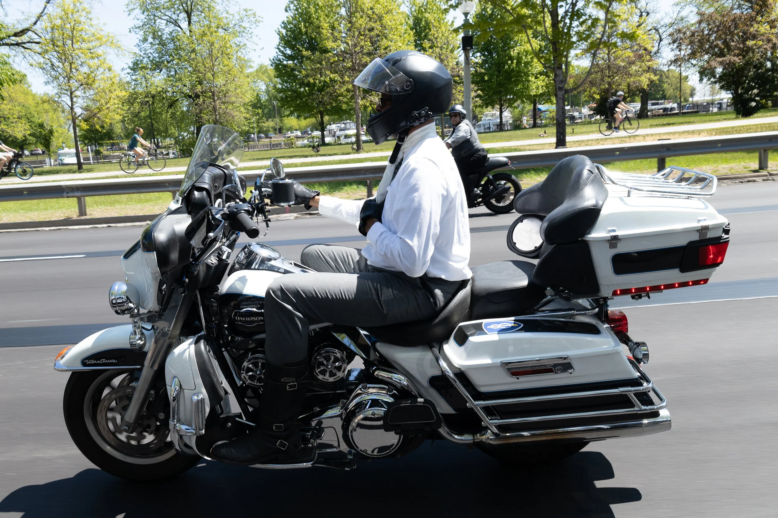 A person riding a white Harley Davidson motorcycle on a city street, wearing a black helmet, white shirt, and gray pants. The motorcycle has a large seat and chrome details. Other cyclists are riding on a bike path in a park with trees and grass in t
