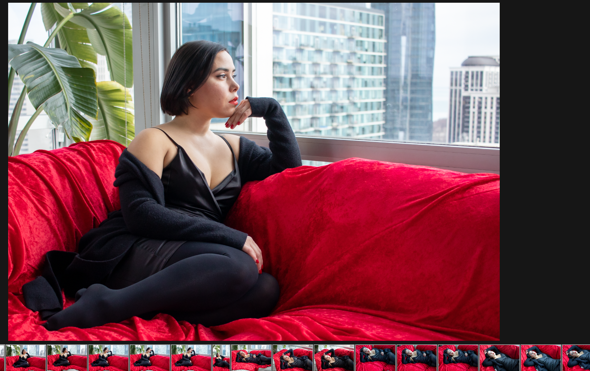 A woman with dark hair and light makeup sitting on a red velvet couch near a large window, looking thoughtfully outside with city buildings in the background. She wears a black satin camisole and a black cardigan.