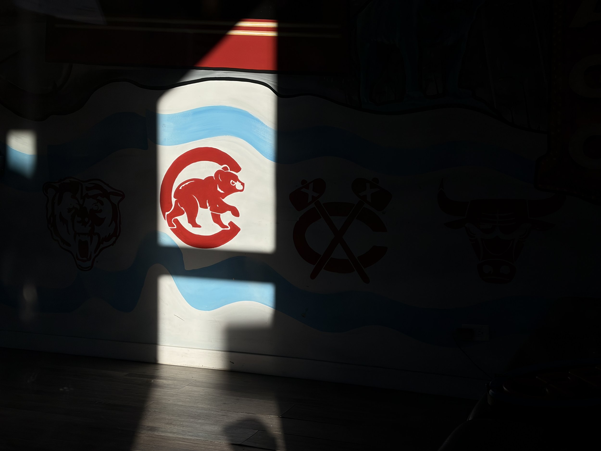 Photo showing a book with a red and white striped cover, illuminated by sunlight, placed on a dark surface with a colorful poster or artwork with various designs and symbols, some featuring animals and tools, in the background.