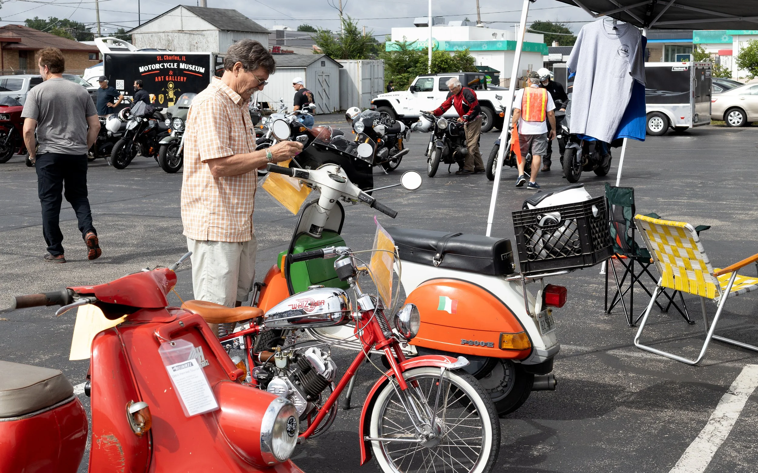 A man in a checkered shirt examining a motorcycle at an outdoor motorcycle event with several motorcycles on display, a tent with t-shirts, and people walking around.