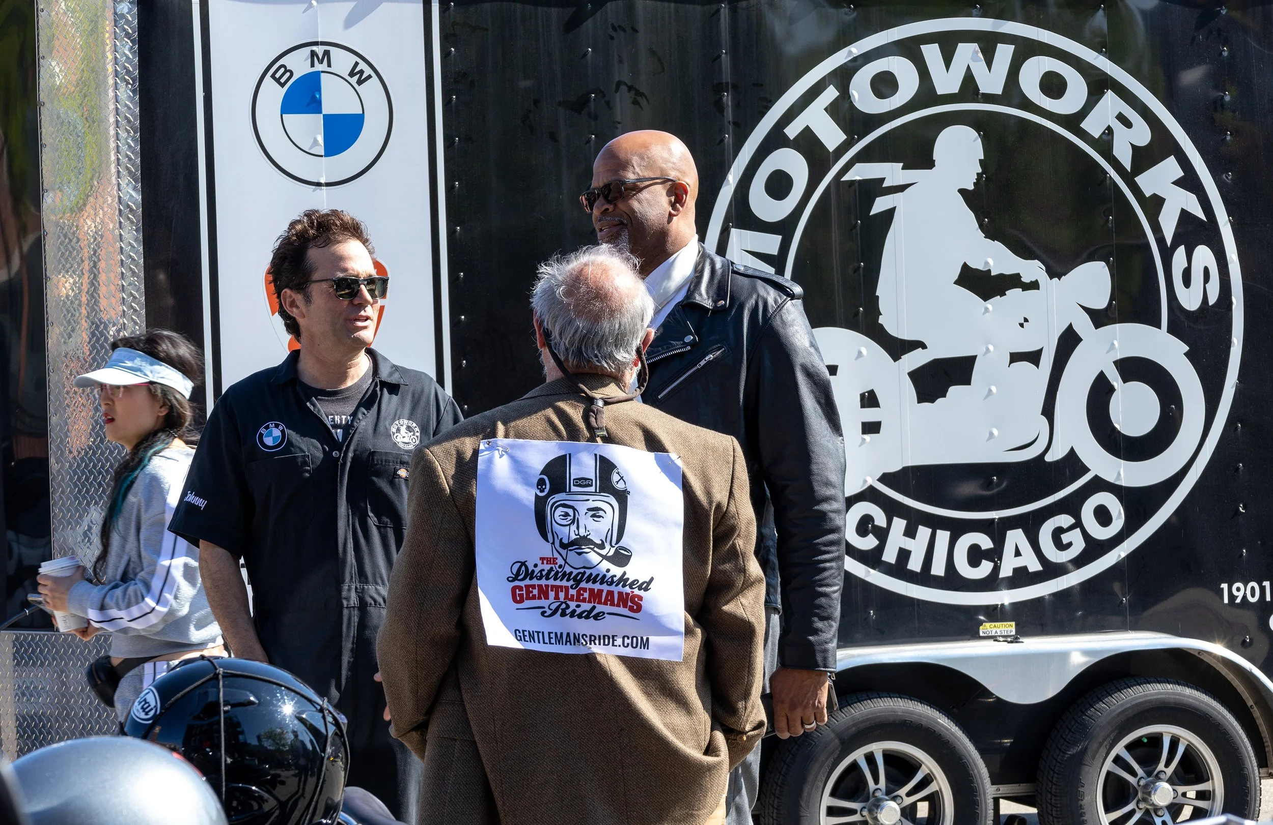 People standing in front of a large truck with the logo for the Chicago BMW Touring Show. The man in the center is wearing sunglasses and talking to an elderly man with a sticker for a ride event on his back, and other people and motorcycles are near