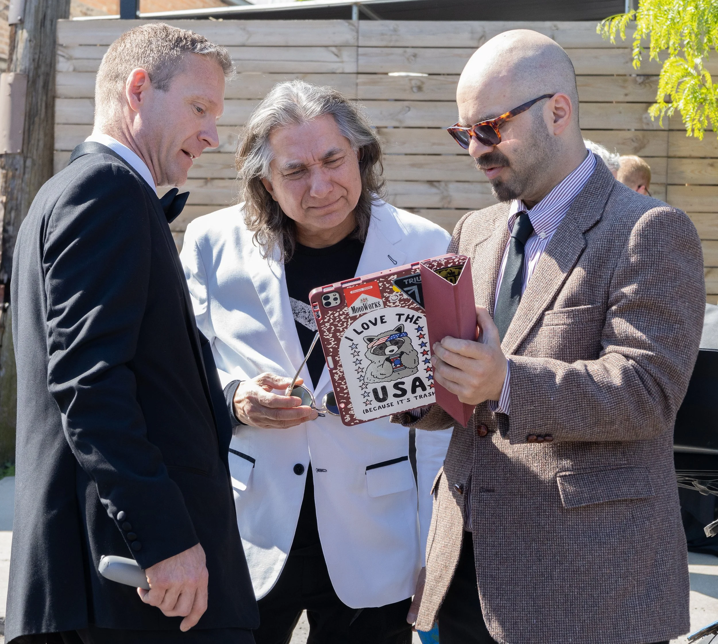 Three men dressed in formal attire, possibly at an outdoor event, looking at a phone together. One man is holding a pink gift bag with stickers and a sticker that reads 'Love the USA'. The background features a wooden fence and some greenery.