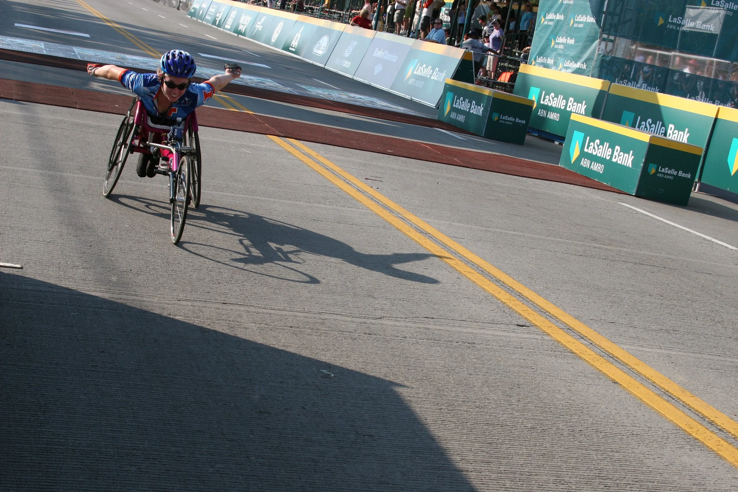 A person in a wheelchair crossing the finish line at a race event, with spectators and banners in the background.