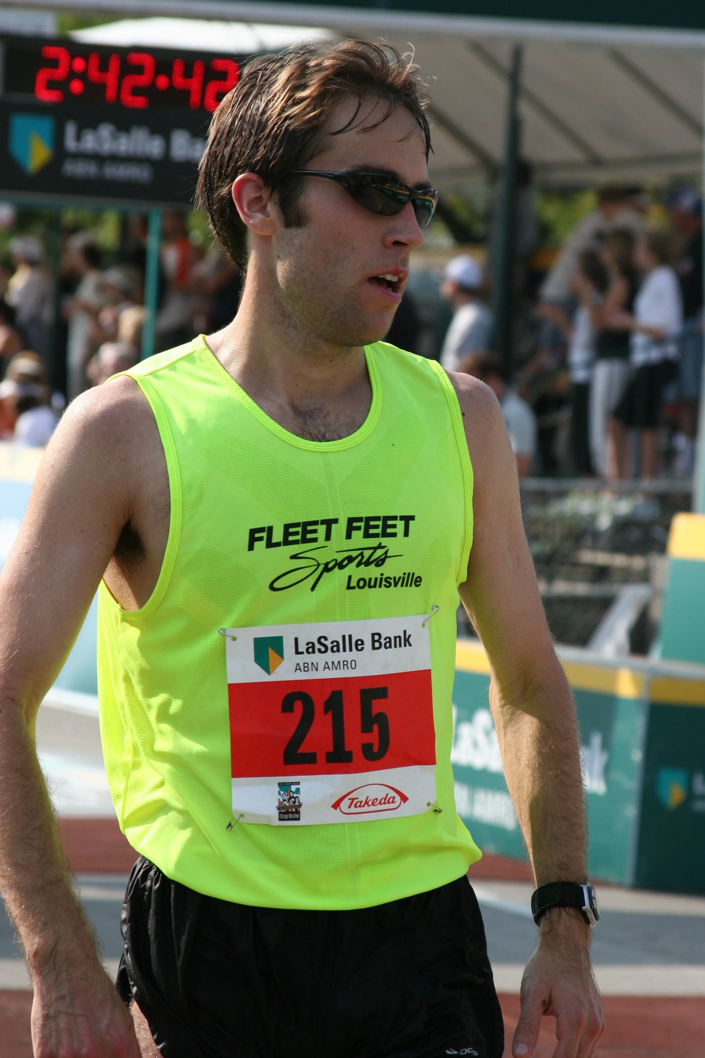 Male runner in a bright yellow tank top with a bib number 215, wearing sunglasses and a black watch, participating in a race at a stadium.