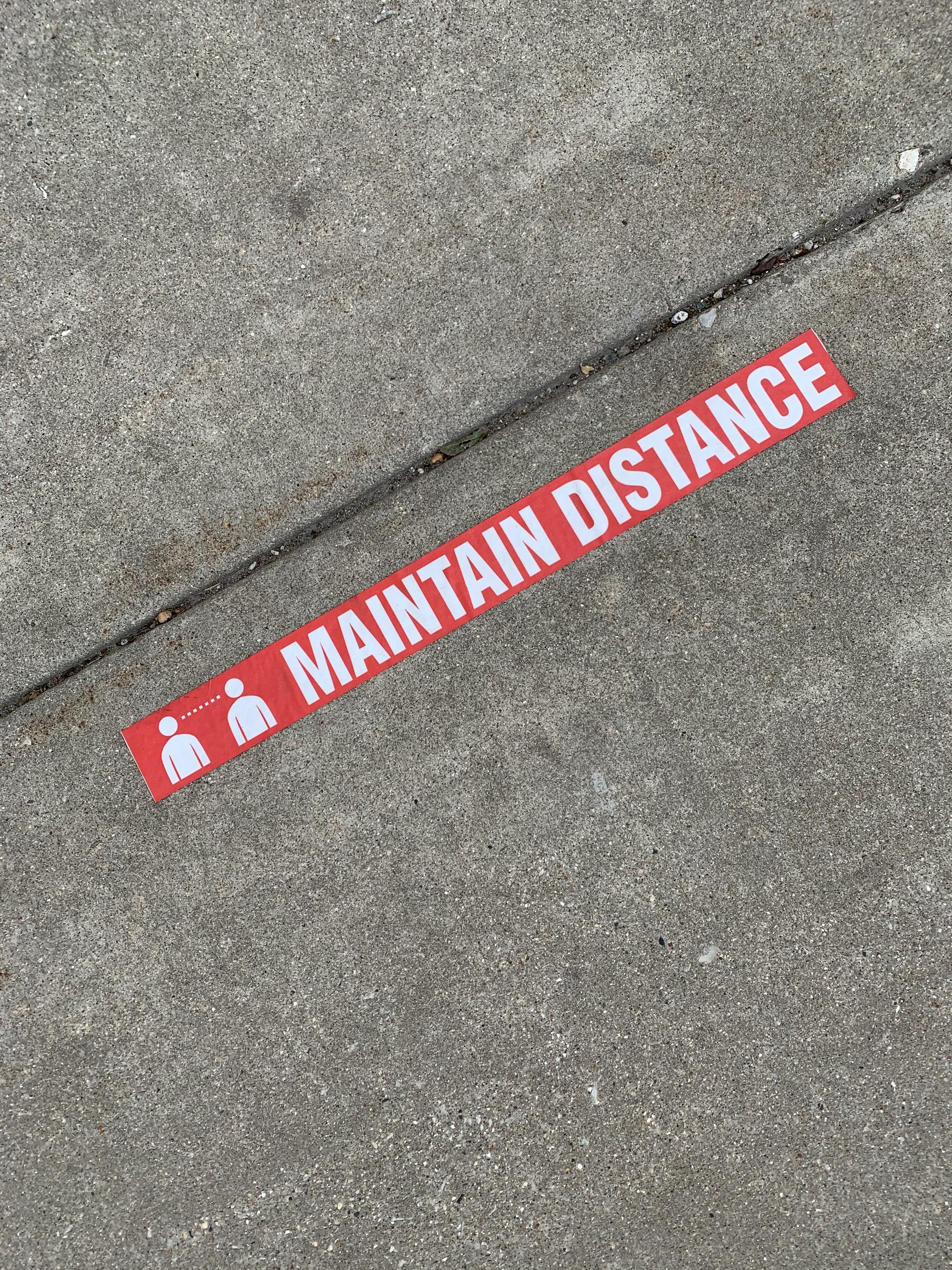 Red tape on the sidewalk with white text and icons, reading "MAINTAIN DISTANCE" and showing two figures separated by dots.
