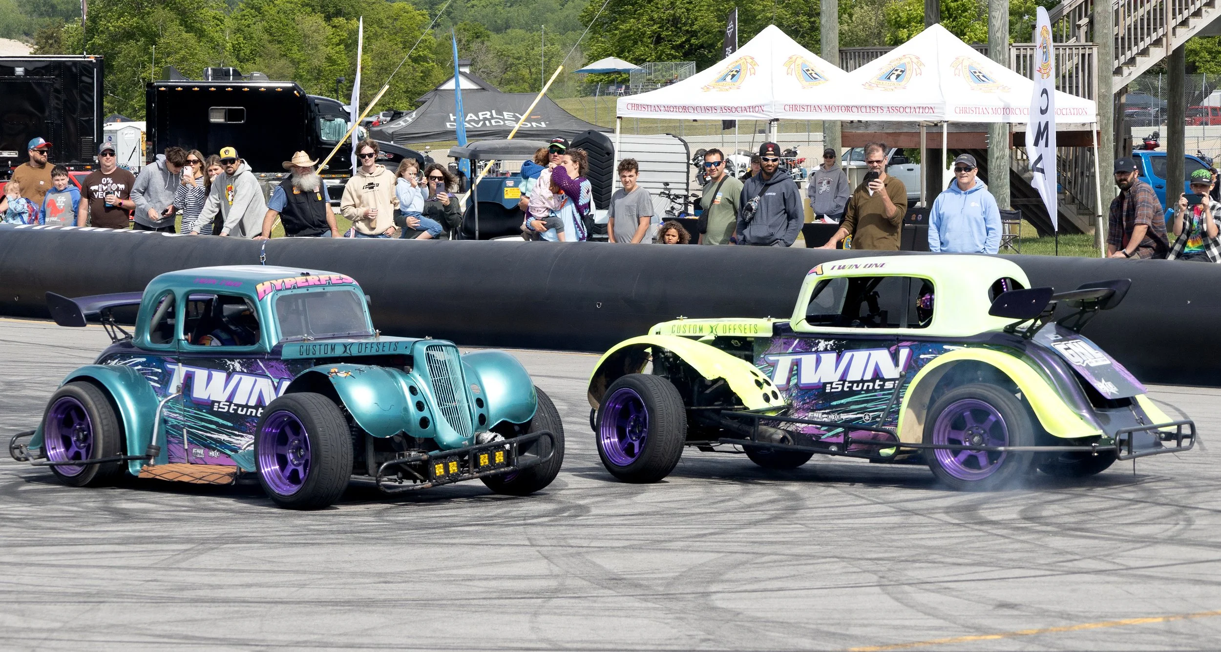 Two colorful stunt race cars on a track with a crowd of spectators watching from the sidelines.