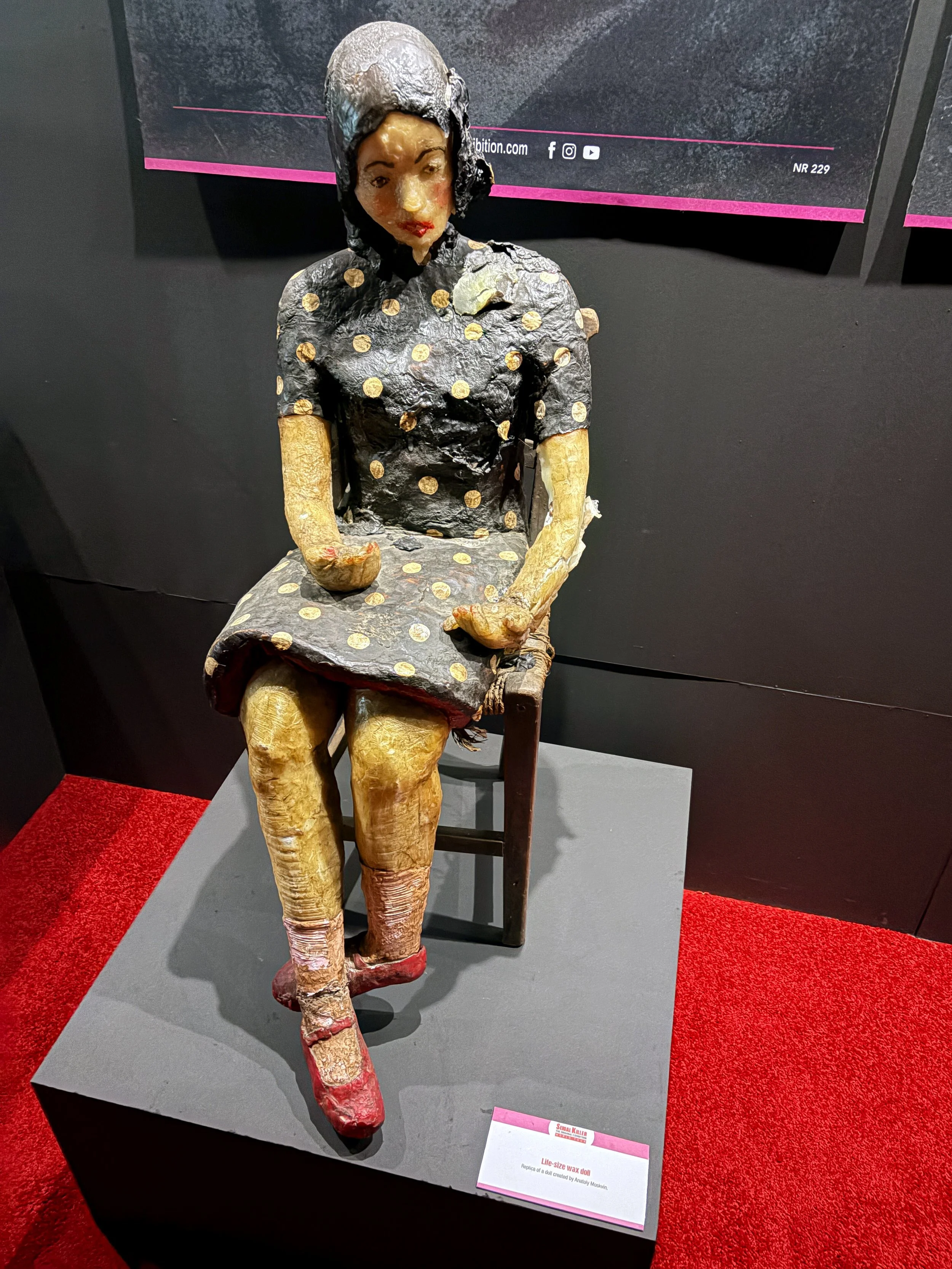A life-size wax doll of a woman sitting on a chair, dressed in a black dress with gold polka dots, with red shoes. The doll has black hair covered with a black and silver hat, a fair complexion, and red lipstick.