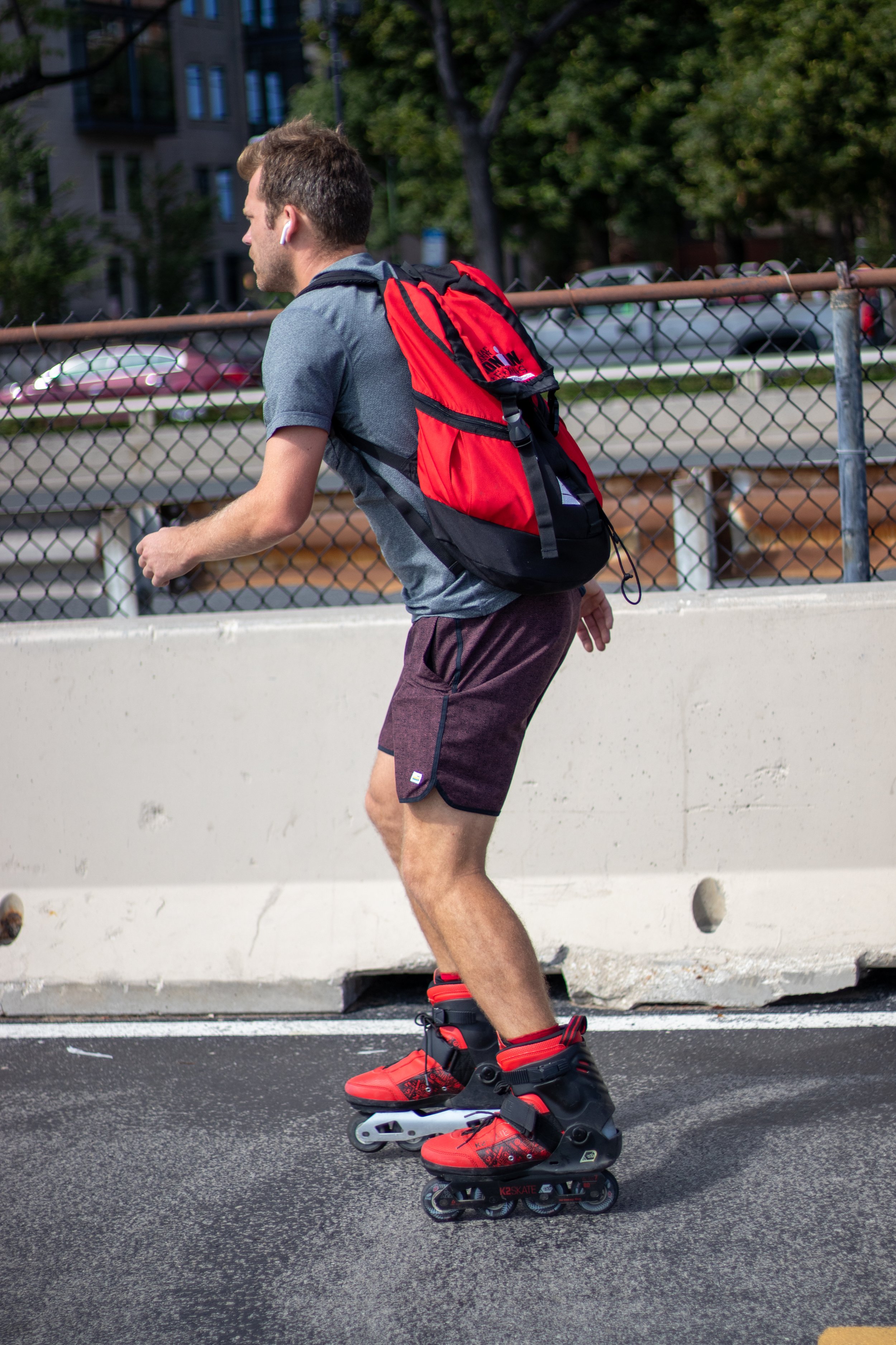 A man wearing a gray t-shirt, dark shorts, red and black rollerblades, and a red backpack is rollerblading on a city street with a concrete barrier and chain-link fence in the background. He has short brown hair and is using AirPods. There are trees 