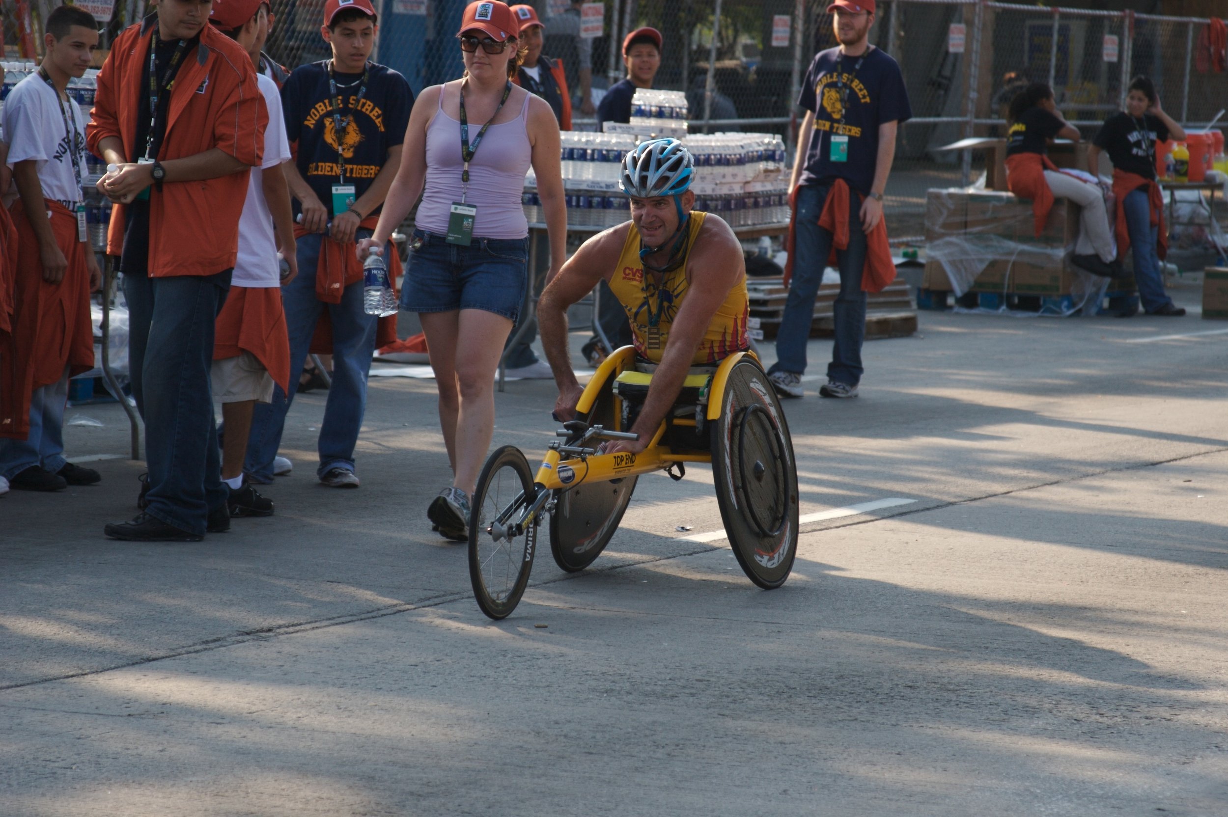 A man in a yellow cycling jersey rides a handcycle with a helmet, while a woman in a purple tank top and blue jeans walks beside him during an outdoor event. Several onlookers, some wearing shirts with the words 'Noble Street Golden Tigers,' are gath