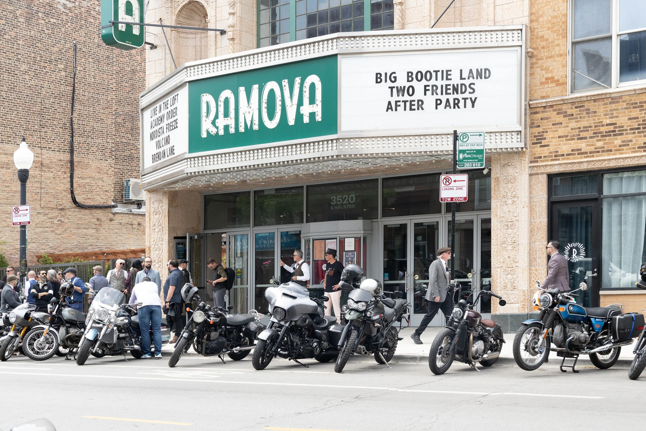 Crowd of people standing near a row of parked motorcycles in front of a building with a marquee sign that says 'RAMOVA'. The building has a brick and stone exterior, large glass windows, and the sidewalk has signs for parking and loading zones.