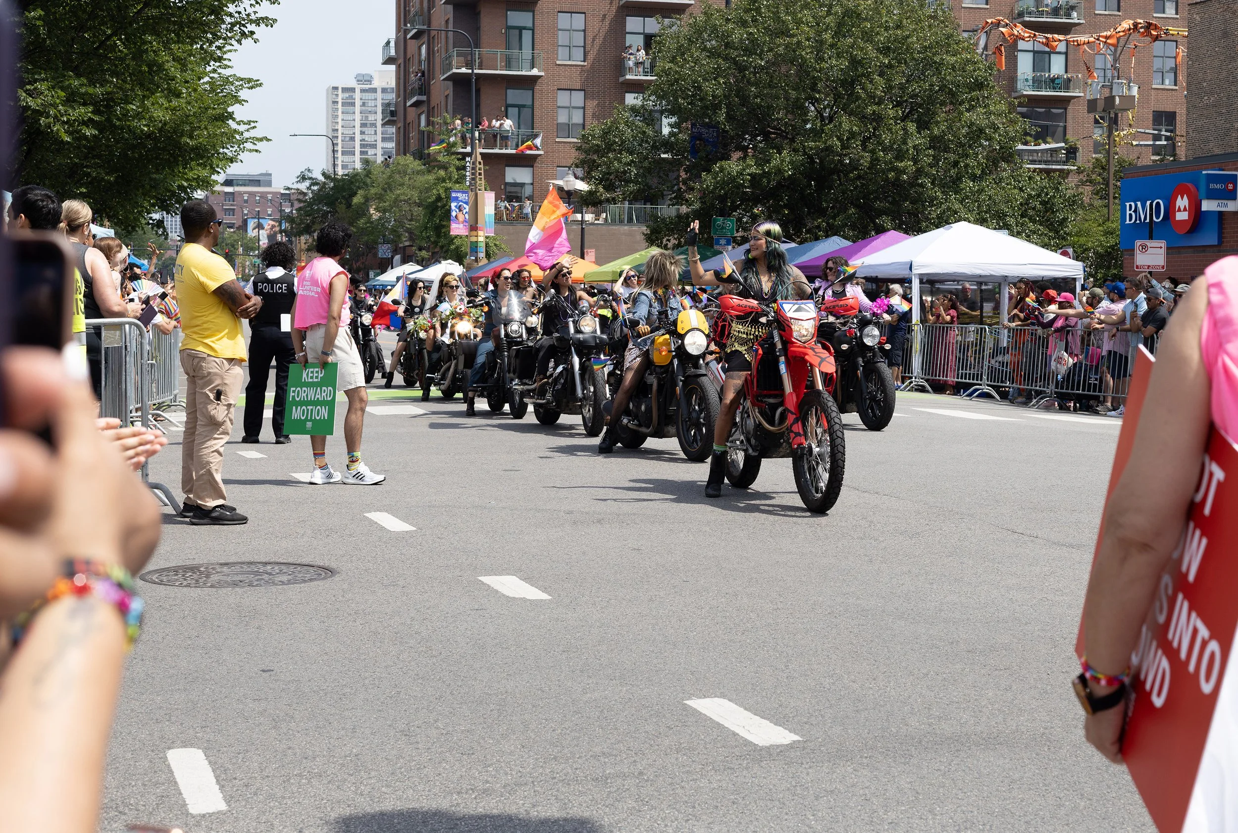 Crowd of people participating in a parade or rally on a city street, with some on motorcycles decorated with rainbow flags and colorful accessories, and spectators standing behind barriers along the sidewalk.