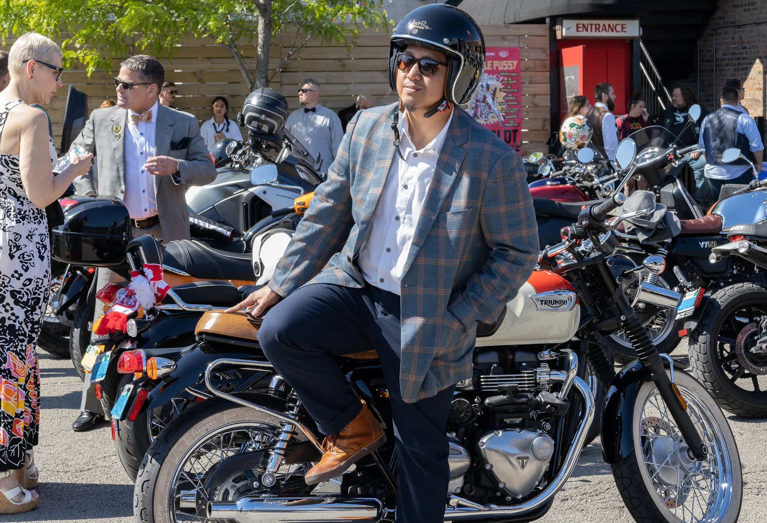 A man wearing sunglasses, a plaid blazer, and a helmet is sitting on a Triumph motorcycle at an outdoor event with several people, including women and men, talking and standing near parked motorcycles, with a building and a sign that says "ENTRANCE" 