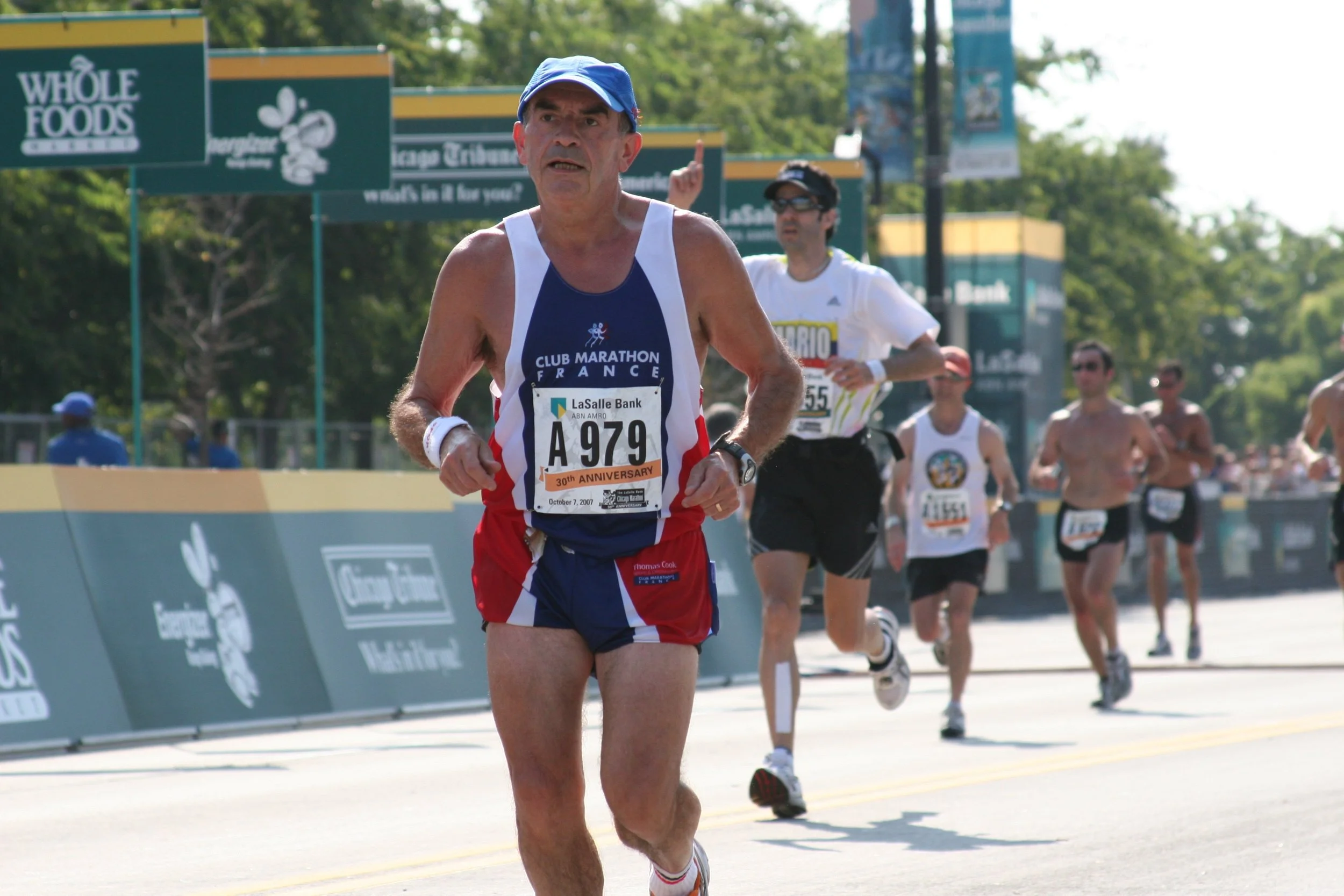 A group of runners competing in a marathon race on a bright, sunny day, with a focus on an older male runner wearing a blue cap, a sleeveless shirt, and red shorts