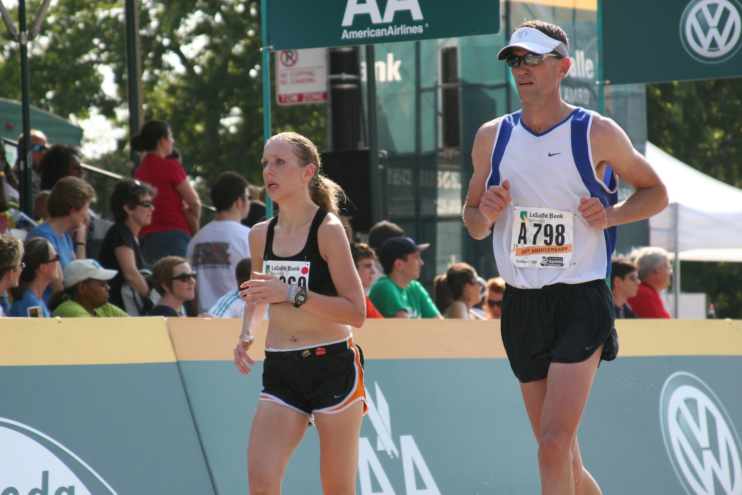 Two marathon runners, a woman wearing black and orange shorts and a man wearing black shorts and sunglasses, running at an outdoor race with spectators watching. There are signs and banners for LaSalle Bank and Volkswagen in the background.