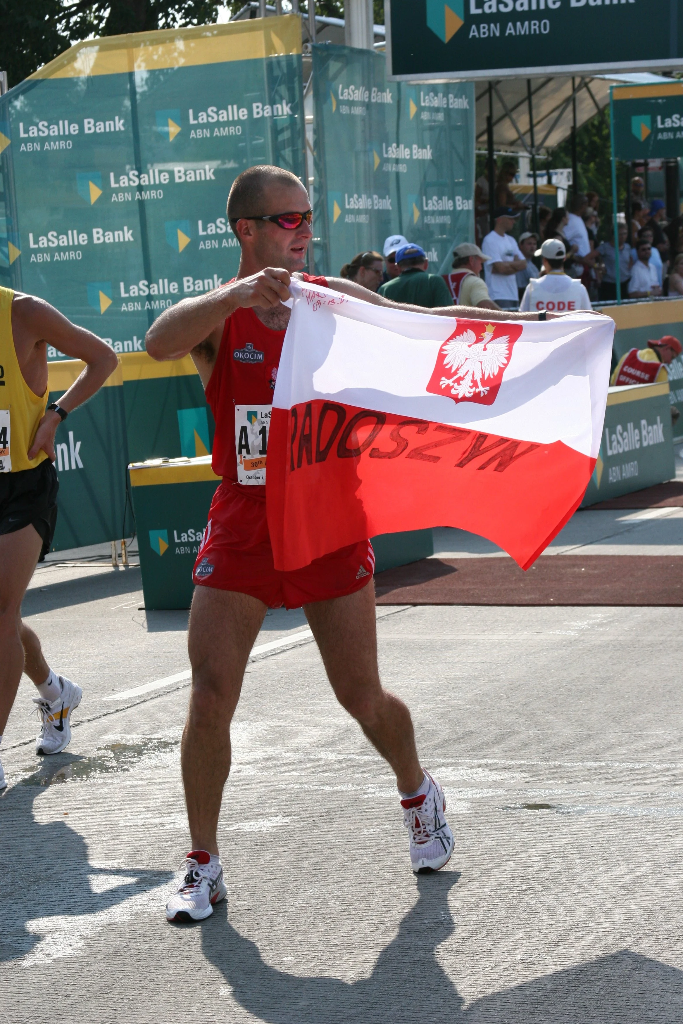 A male marathon runner holding a Polish flag at a race event, with spectators and sponsor banners in the background.
