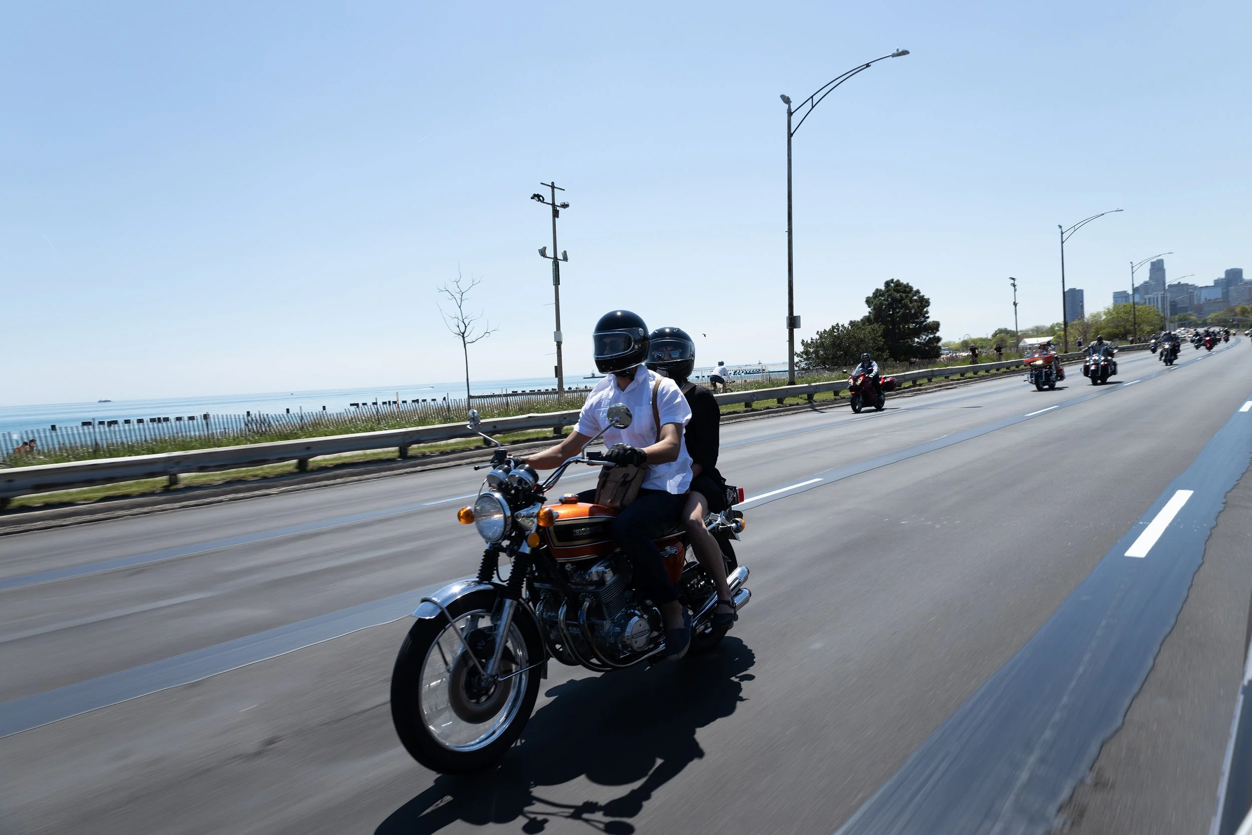 Motorcyclists wearing helmets ride along a city highway near the coast, with city skyline and blue sky in the background.