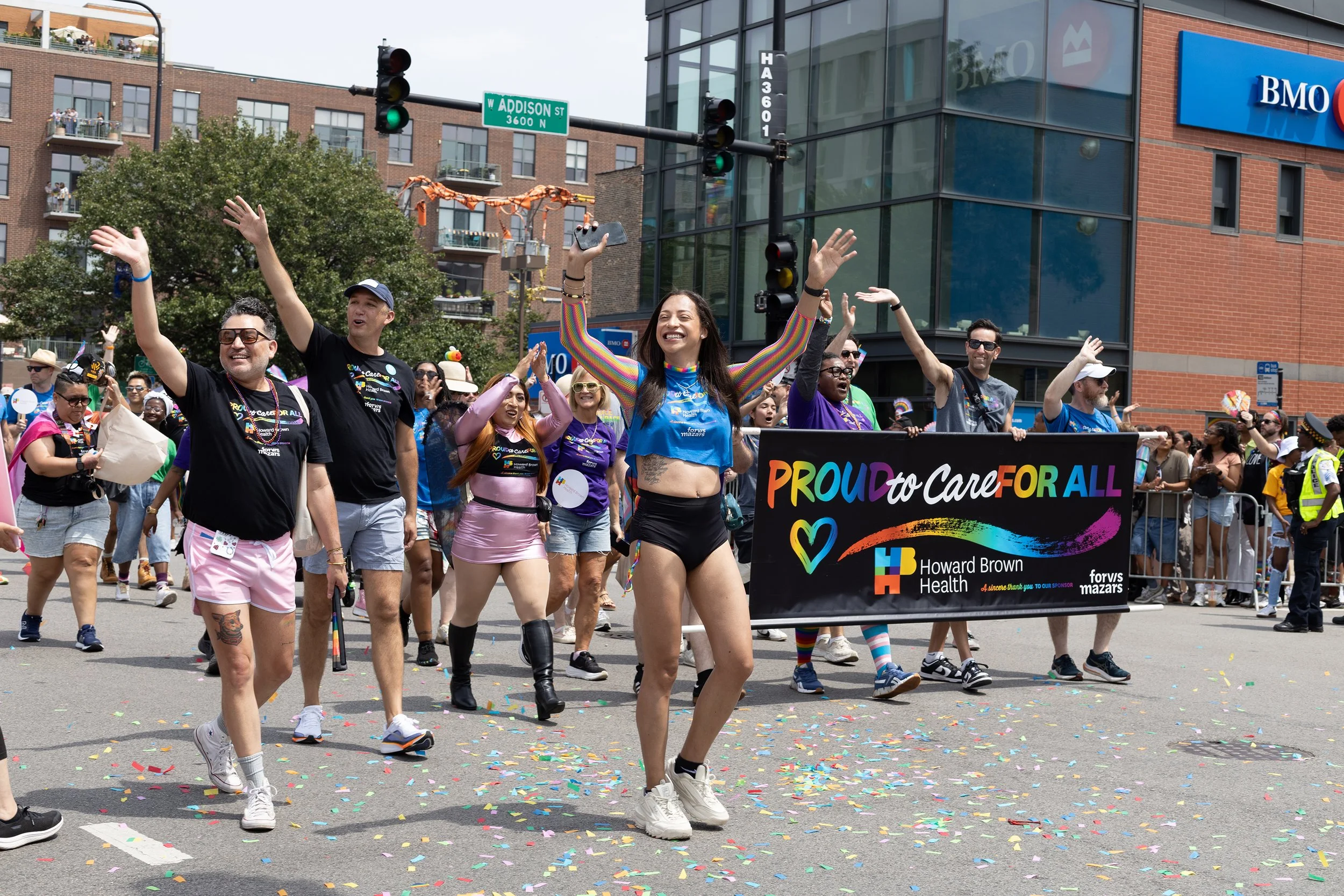 Group of diverse people participating in a pride parade, holding a rainbow-colored banner that says "Proud to Care for All," with some smiling and waving, colorful confetti on the street, and a city building in the background.