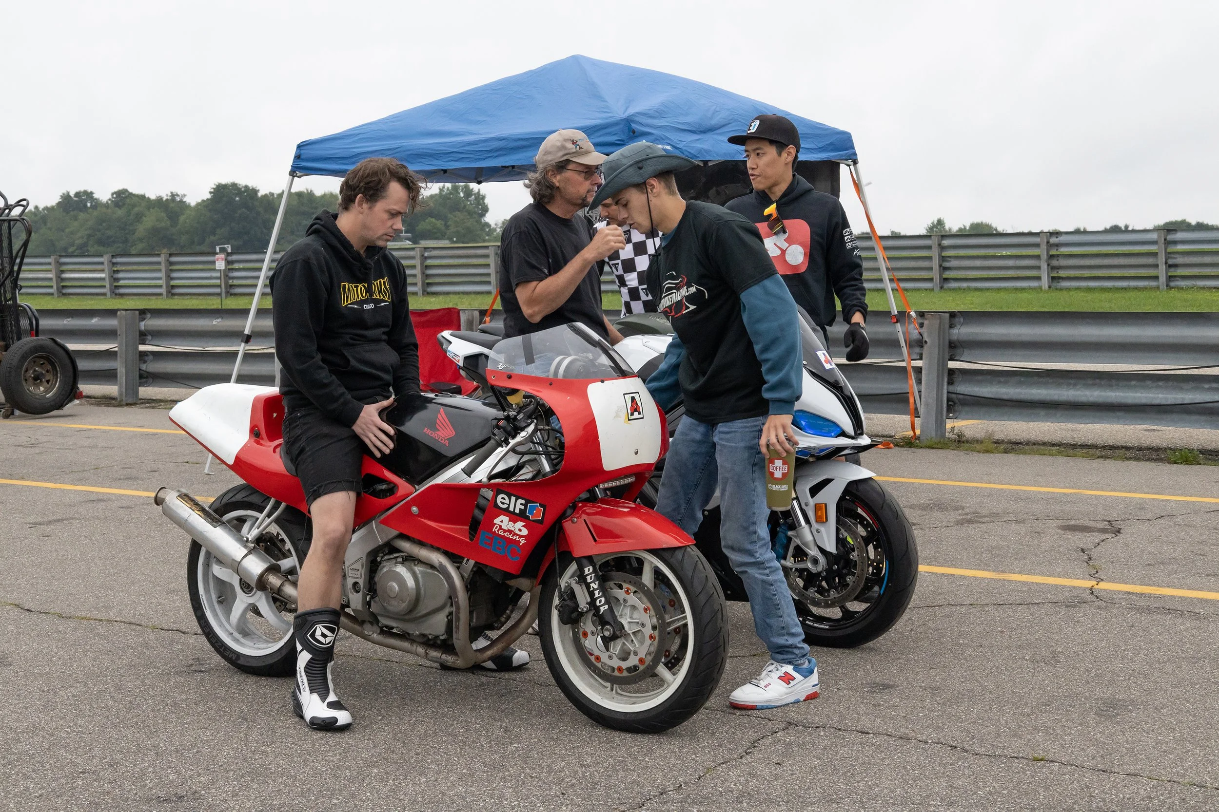 A group of five men gathered around a red and white racing motorcycle on a track, with a blue canopy tent in the background. They appear to be inspecting or discussing the motorcycle.