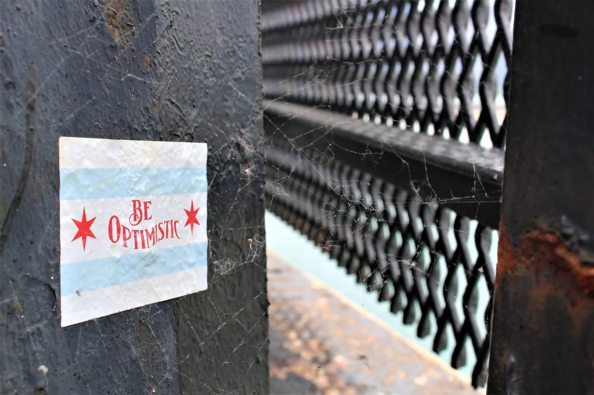 A close-up of a dark gray concrete post with cobwebs and a partially visible sticker that reads "BE OPTIMISTIC" with red stars. Behind the post, there's a black metal fence with a repeating diamond pattern.