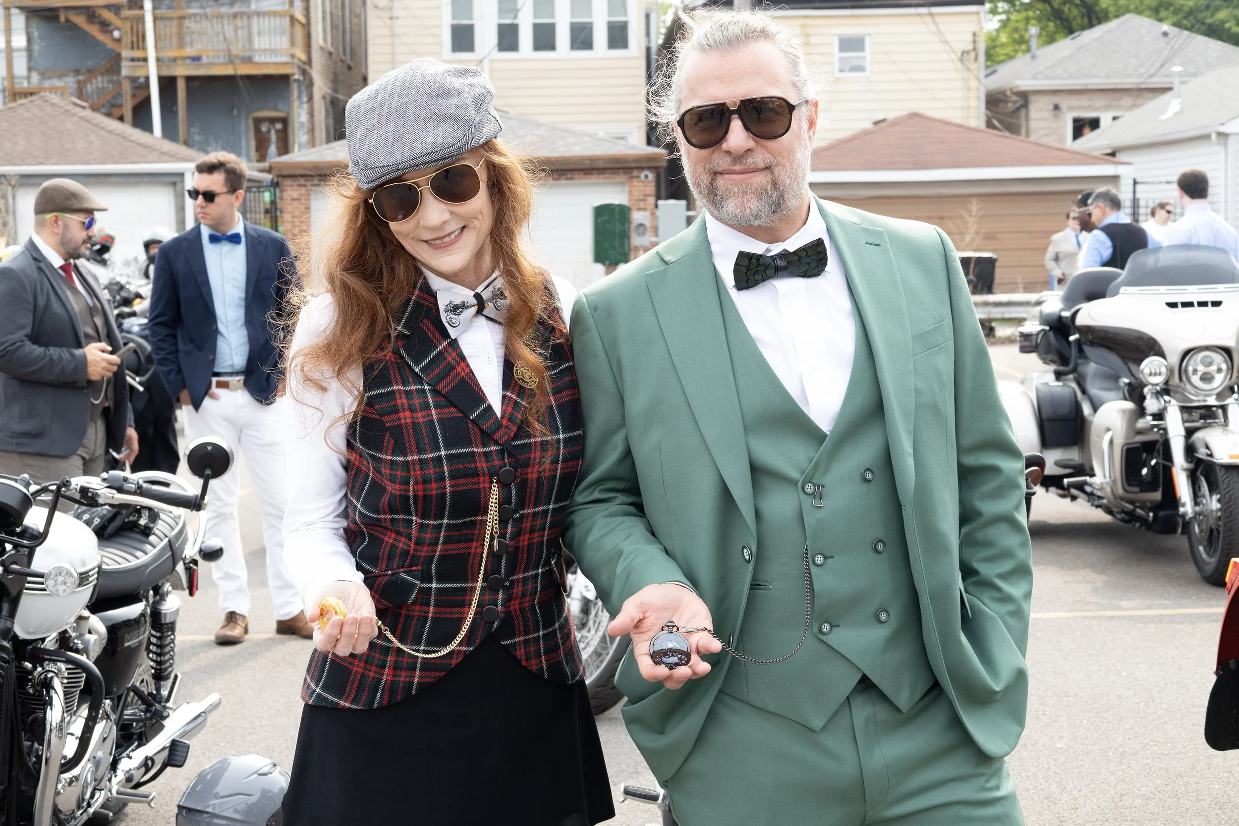 Two people, a woman with long red hair wearing sunglasses, a gray newsboy cap, a white shirt with a bow tie, and a plaid vest, and a man with gray hair and beard wearing sunglasses and a green suit, are holding pocket watches connected by a chain in 