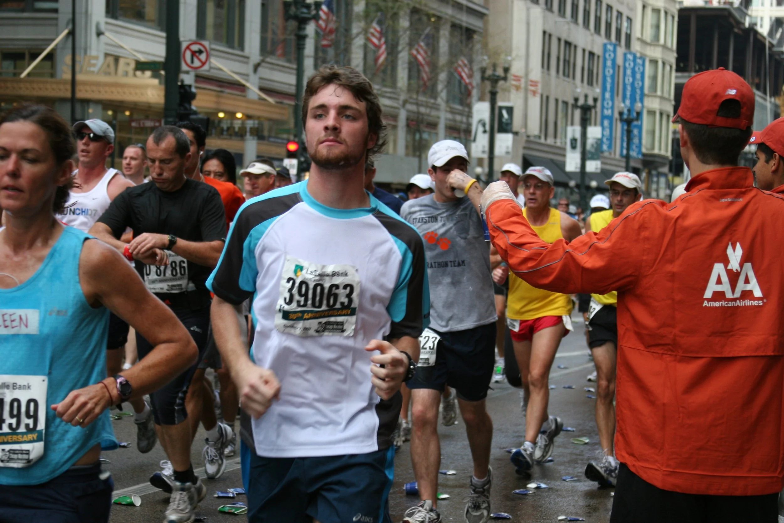 A group of marathon runners participating in a race on a city street, with some individuals wearing numbered bibs and athletic gear. An official in an orange jacket is seen handing out water or aid to the runners.