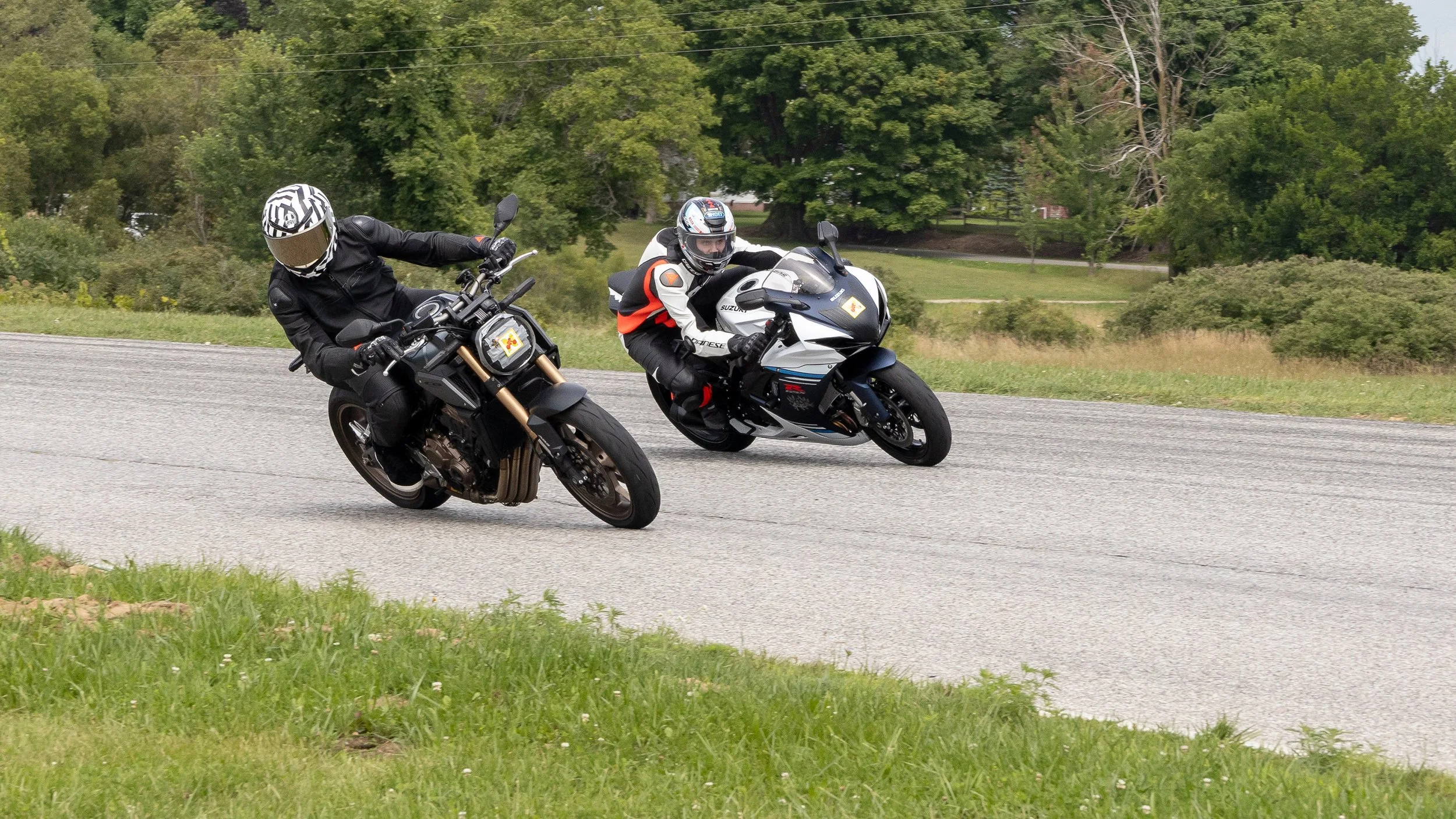 Two motorcyclists leaning into a turn on a rural road with green trees and grass in the background.