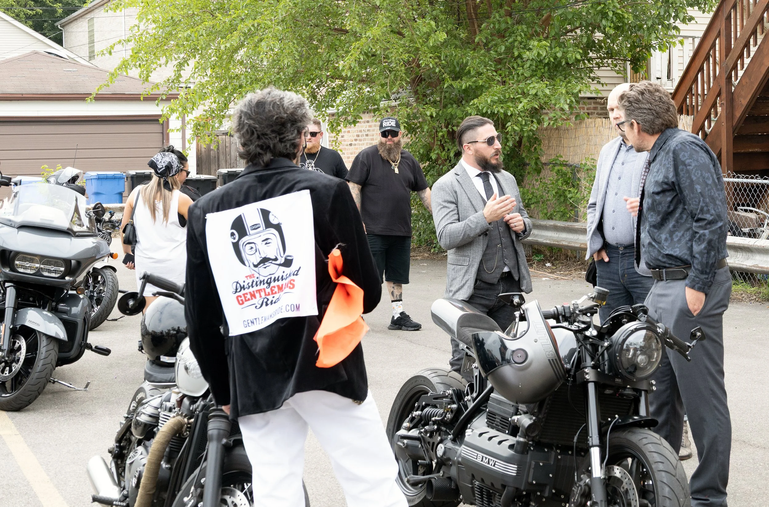 Group of men and women gathered outdoors near motorcycles, engaged in conversation during daytime.