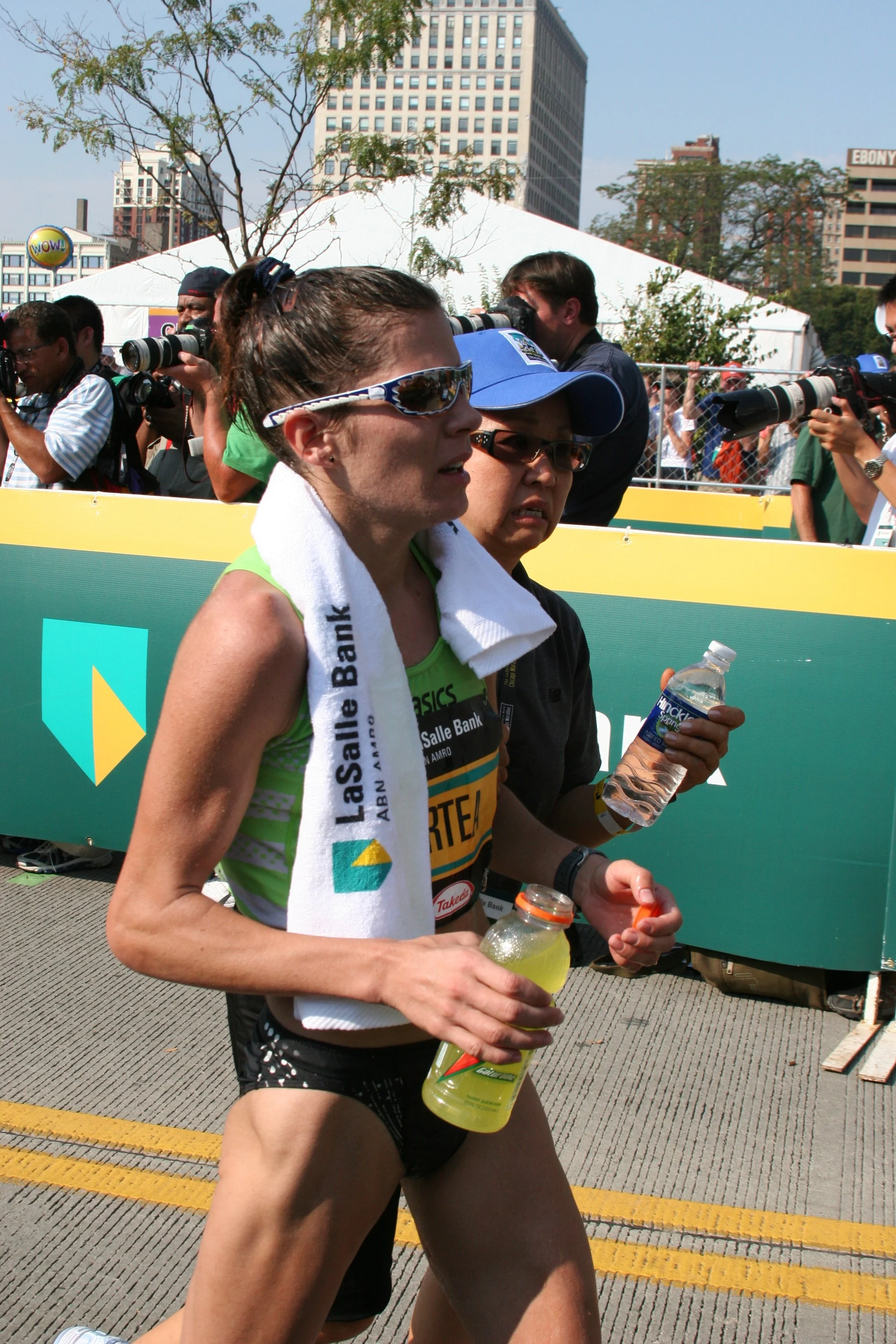 A female marathon runner in a green tank top, black shorts, and sunglasses, holding water bottles, participating in a race outdoors with spectators and photographers behind a fence in an urban setting.
