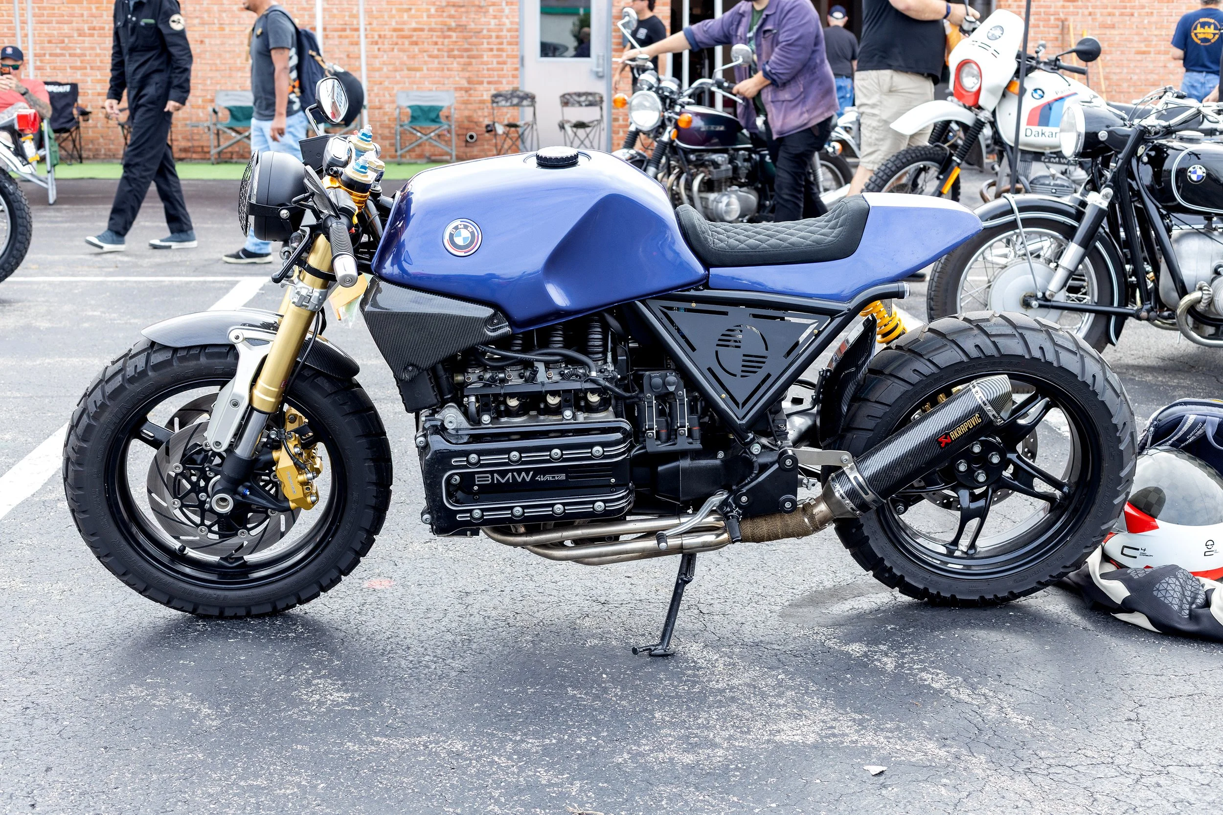 Blue BMW motorcycle with black seat and gold front suspension, parked in a crowded lot at a motorcycle event, with several other motorcycles and people in the background.