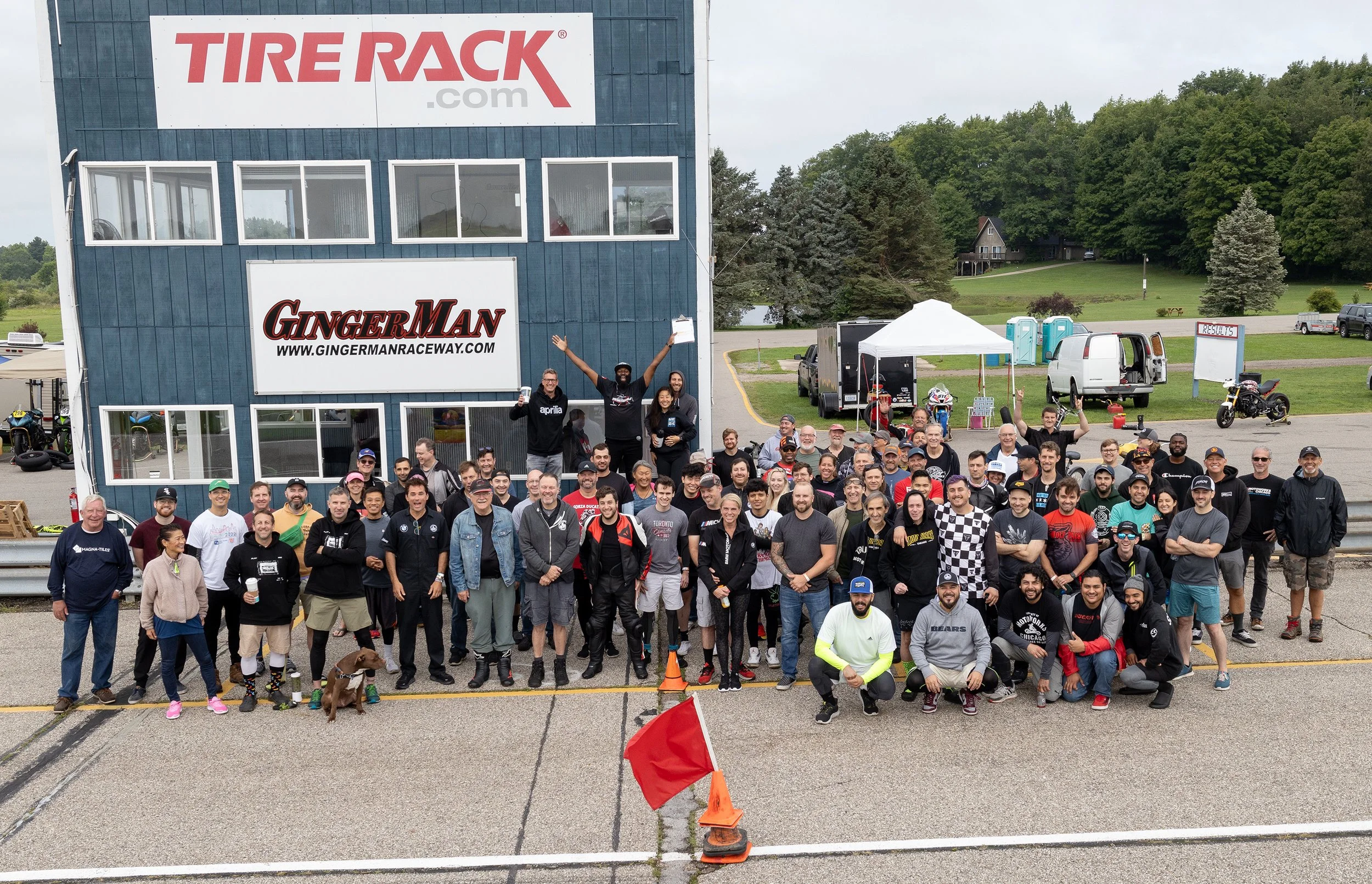 A large group of people gathered outdoors for a group photo at Gingerman Raceway, with a building displaying the TIRE RACK and GINGER MAN logos in the background. The group appears happy, with some individuals raising their hands or holding drinks, a